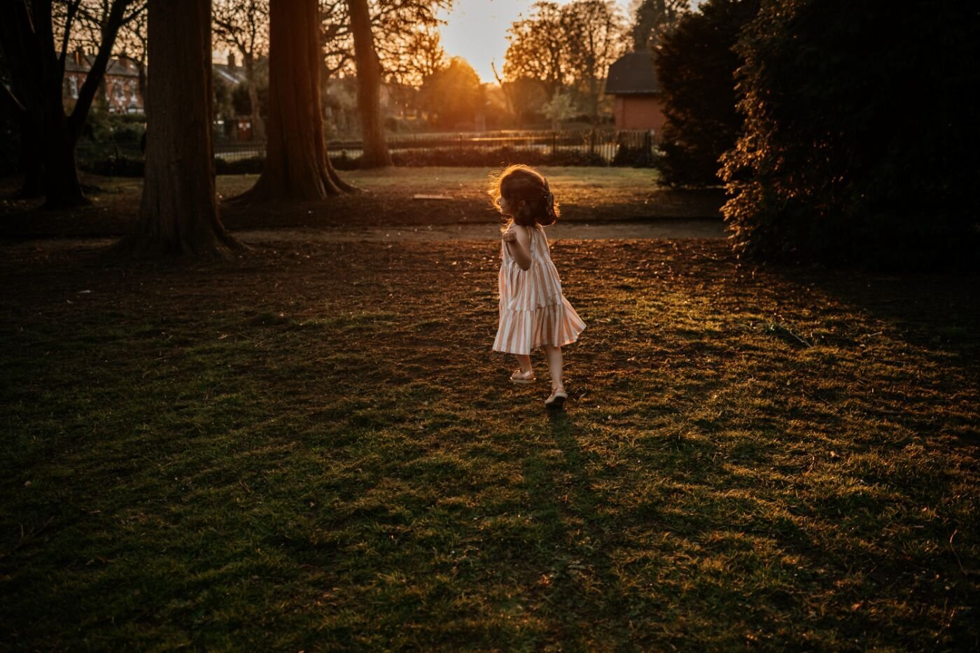 A young girl in a light-colored dress walks away from the camera across a grassy park at sunset, bathed in warm golden light filtering through the trees