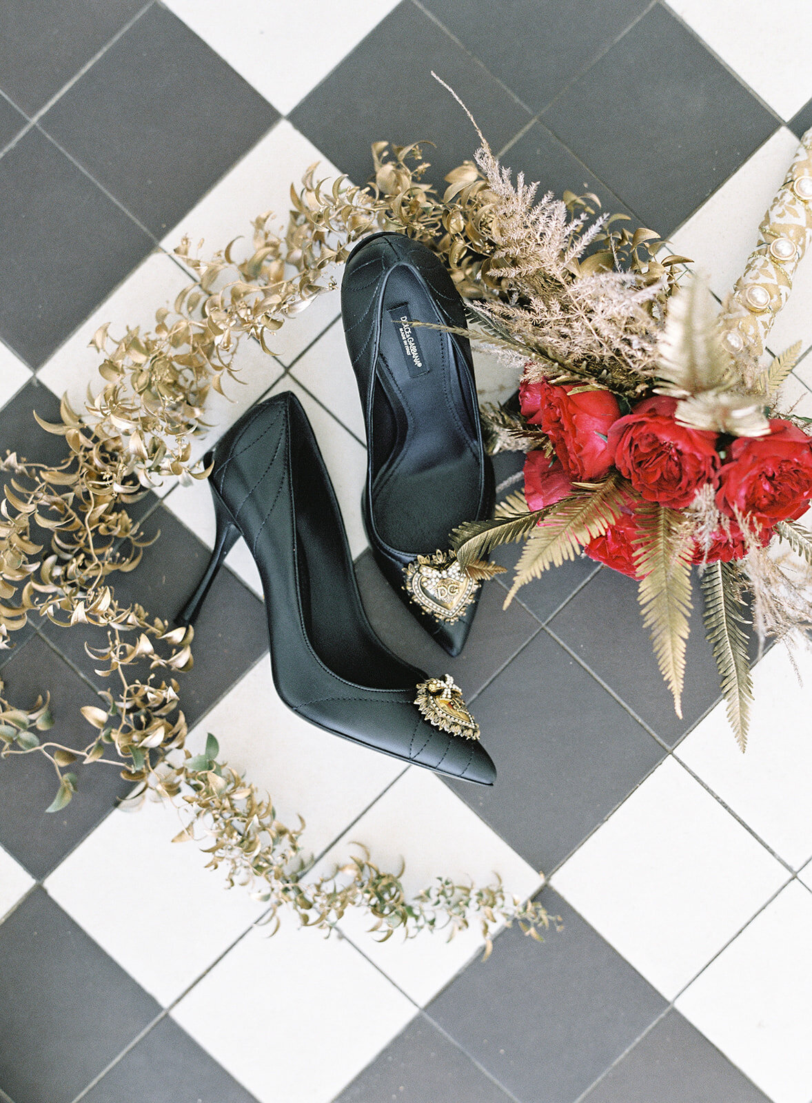 Dolce & Gabbana black bridal shoes with bride's red garden rose and painted gold leaves circling around them. On black and white diamond flooring. Photographed by wedding photographers in Charleston Amy Mulder Photography.