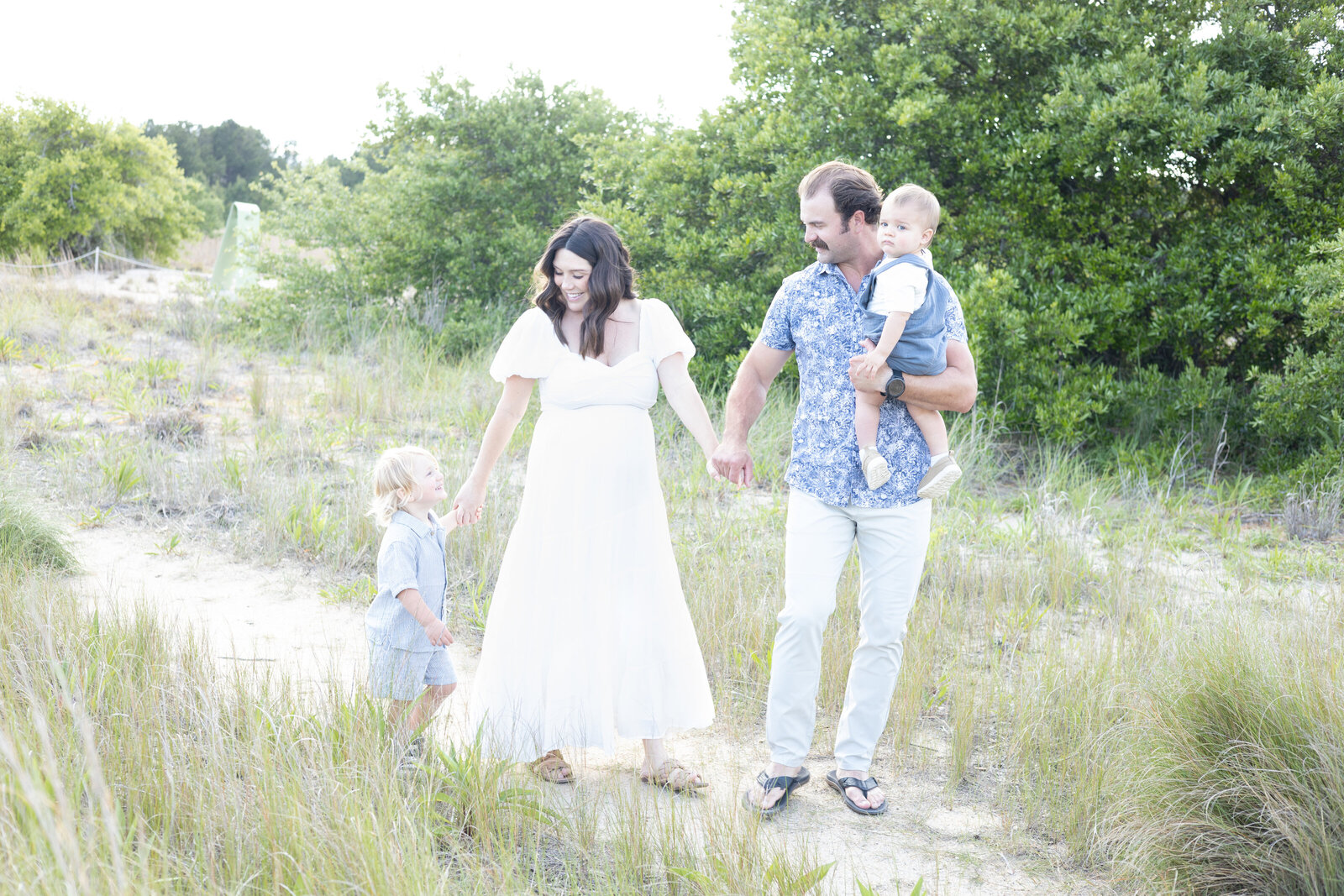 Photograph of a Family of 4 with a pregnant mother in a white dress walking with her husband and 2 toddler sons along a beach trail Virginia Beach Maternity Photography  by Mary Eleanor