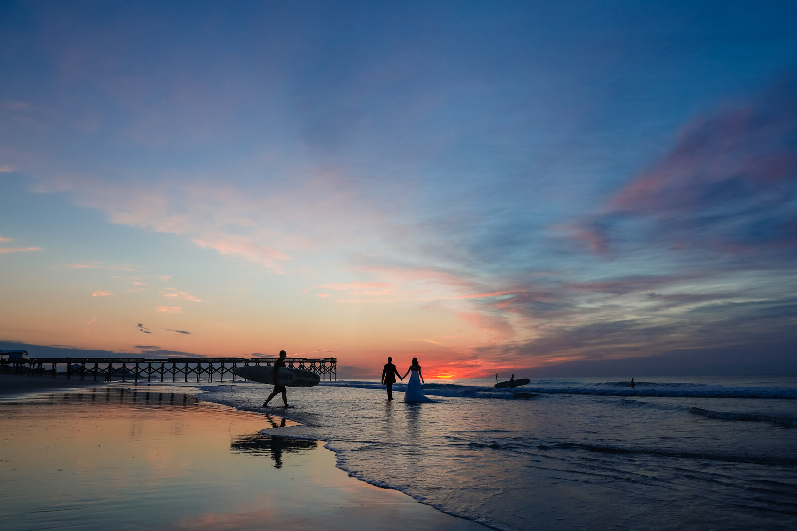 "Newly married couple walks between surfers during sunrise at Crystal pier on Wrightsville Beach North Carolina"