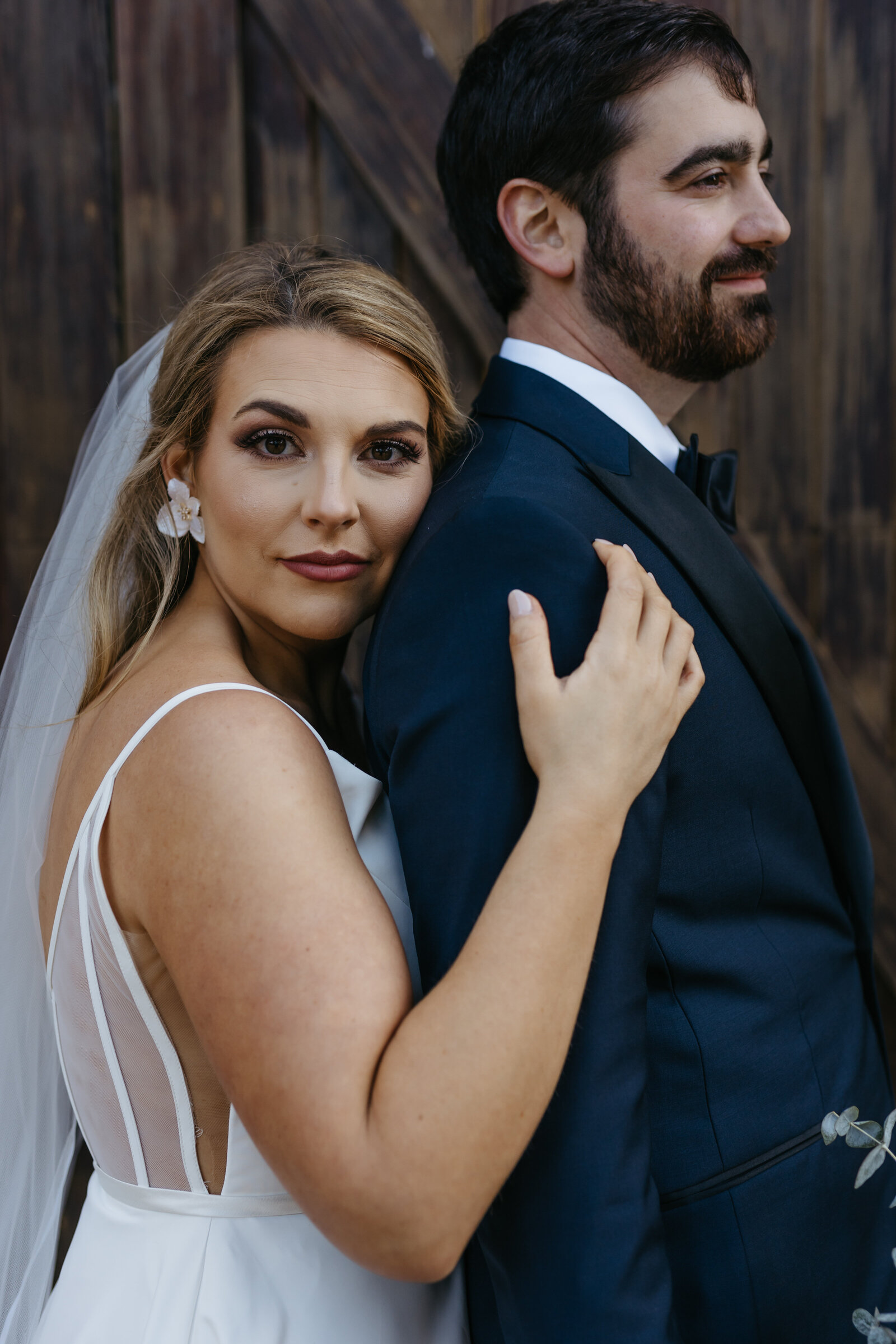 A bride with a long veil leans into a groom's shoulder