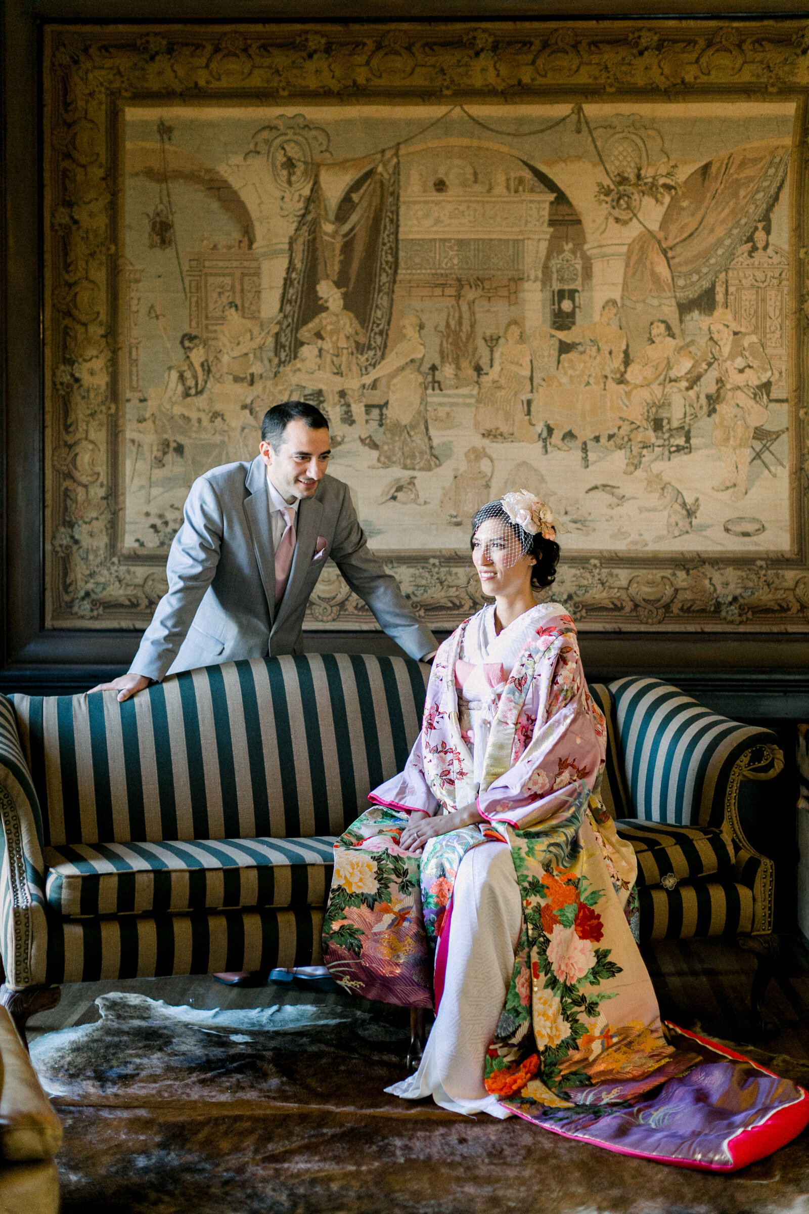 Bride in pastel pink uchikake kimono and groom in light grey suit posing in front of grand tapestry at The Ebell Club Long Beach, available for lovely kimono rental in Los Angeles | ロサンゼルスThe Ebell Club Long Beachの大きなタペストリー前で淡色打掛花嫁と新郎の着物レンタル和婚フォト
