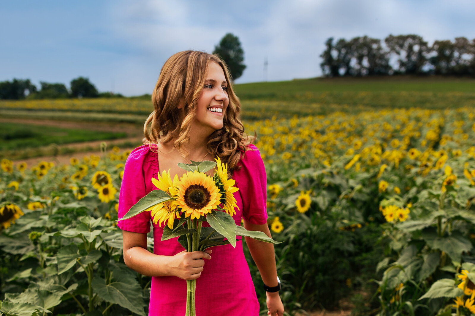 Cute Brown haired girl holds a thick bouquet of sunflowers and wears a bright pink dress for her senior Photo.