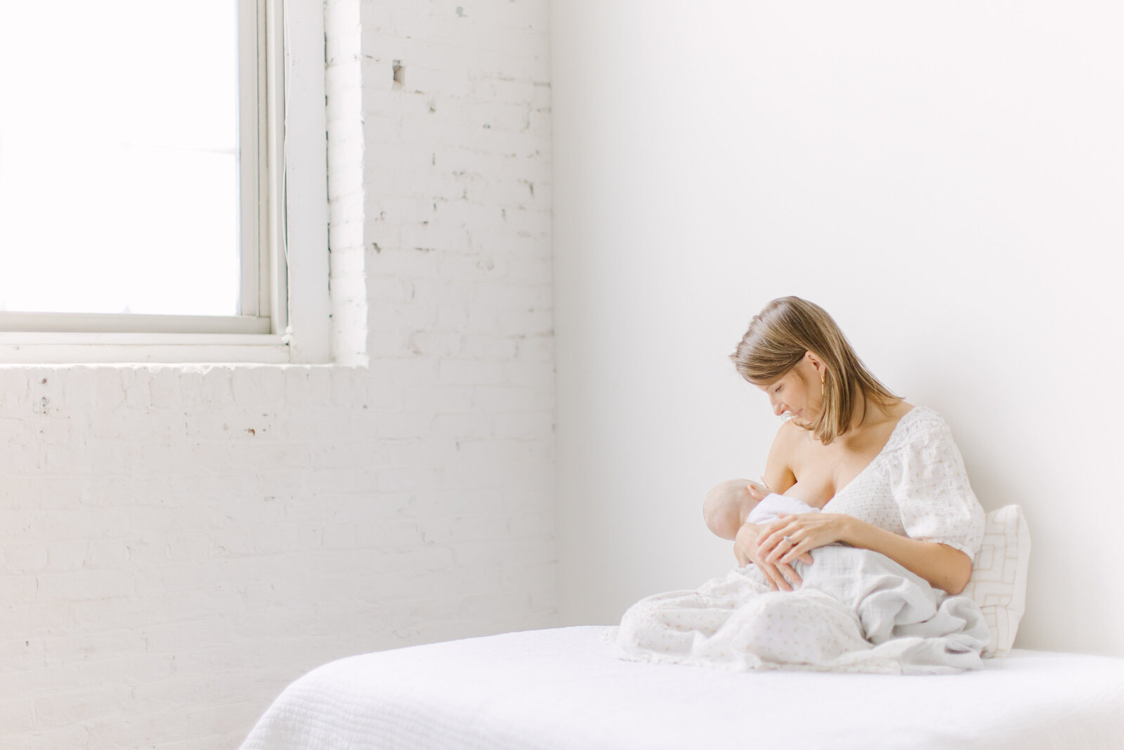 A new mother breastfeeding her baby while sitting on a white bed in a photo studio