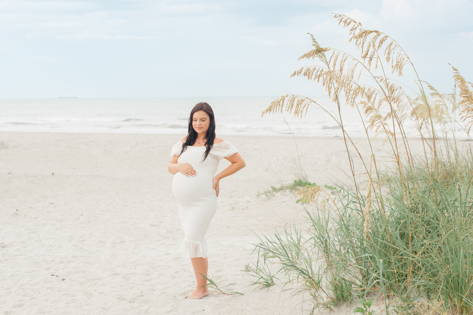 Expectant mom holds her belly with beautiful tall dune grass and the water in the background