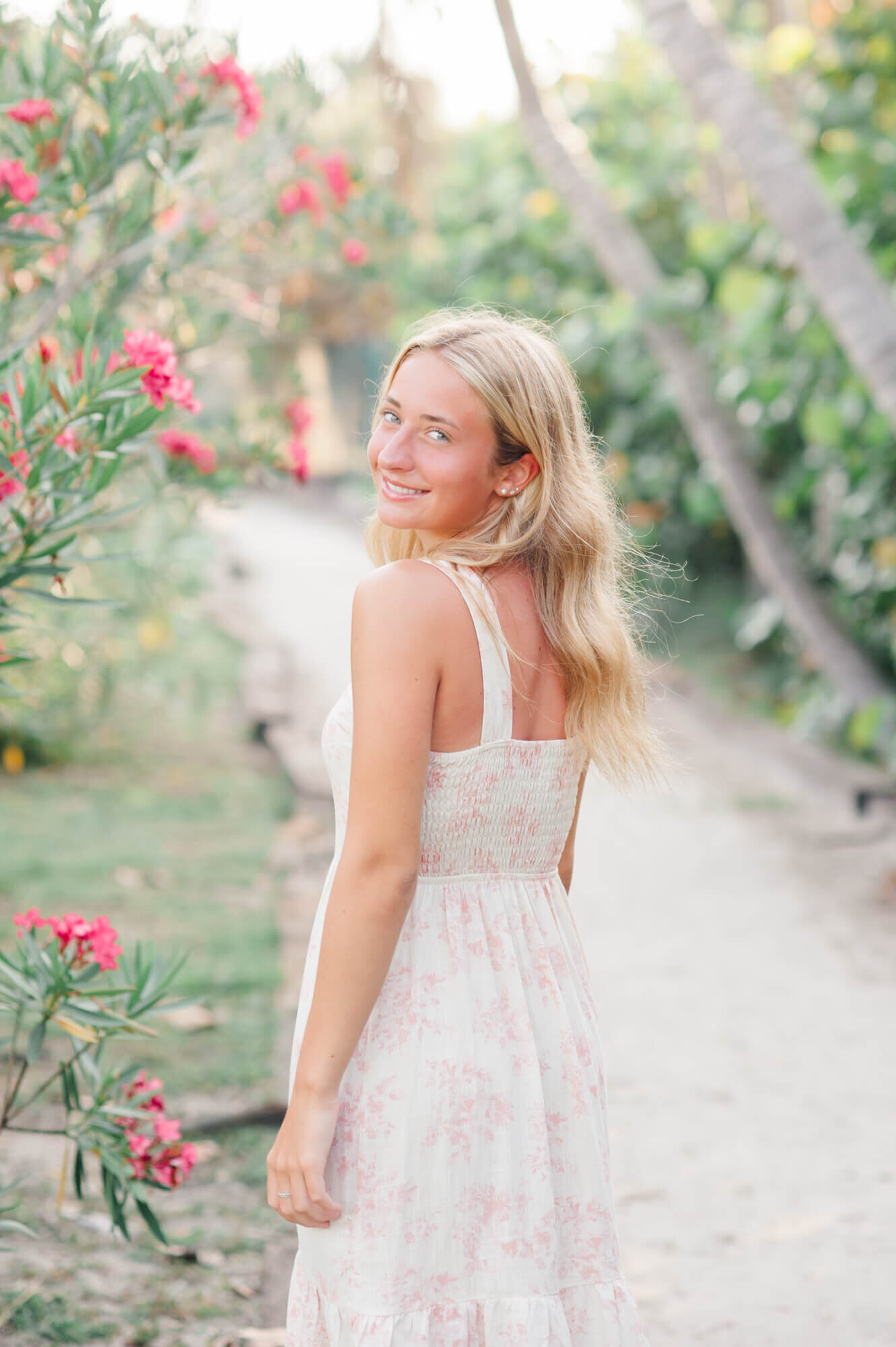 Blonde senior girl stands on beach path next to pink florals and smiles back over her shoulder during her Orlando senior photographer session