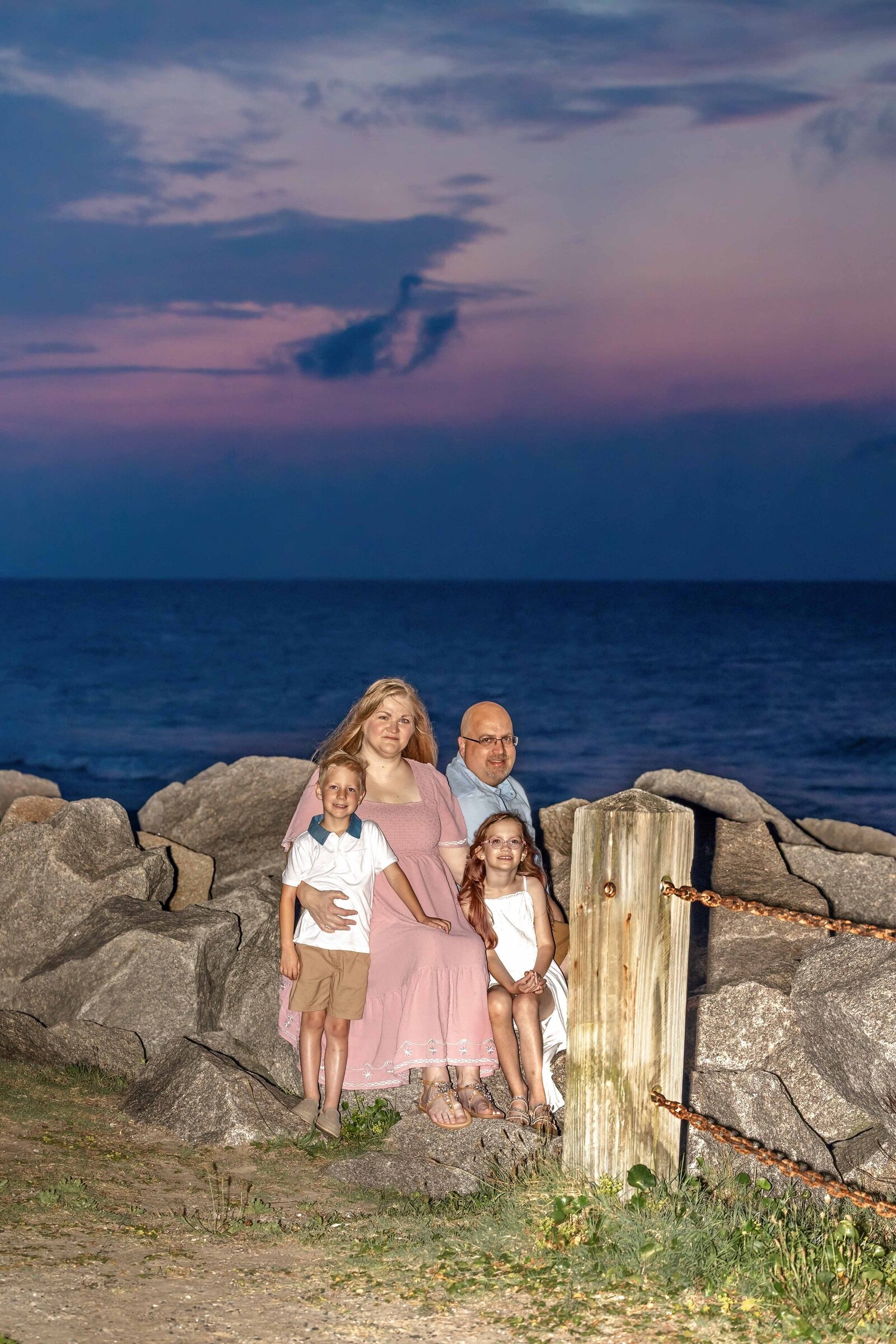 A family of four sits on rocks by the ocean at dusk, dressed in pastel outfits. The deep blue sky and waves create a serene, intimate setting, perfect for artistic family photography in Wilmington, NC, highlighting peaceful coastal moments.