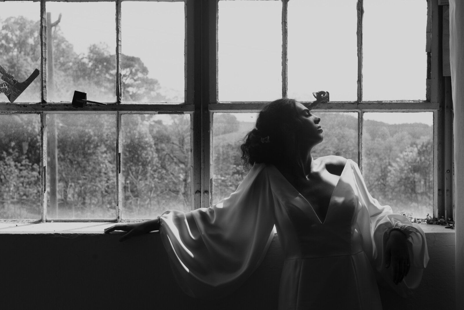 Black and white editorial bridal portrait of a modern bride in a satin gown, posed in dramatic natural light by a vintage window in South Carolina.