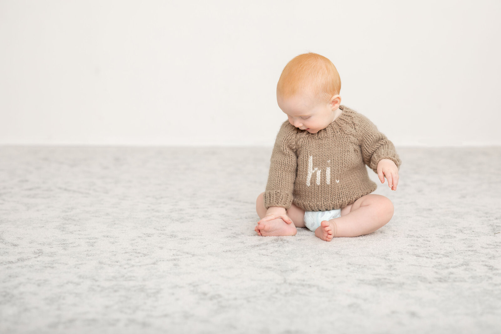 Toddler wearing a brown sweater with the word 'hi' sits on the floor, smiling softly. Captured by Bucks County Photographer Ivory Tree Portraits