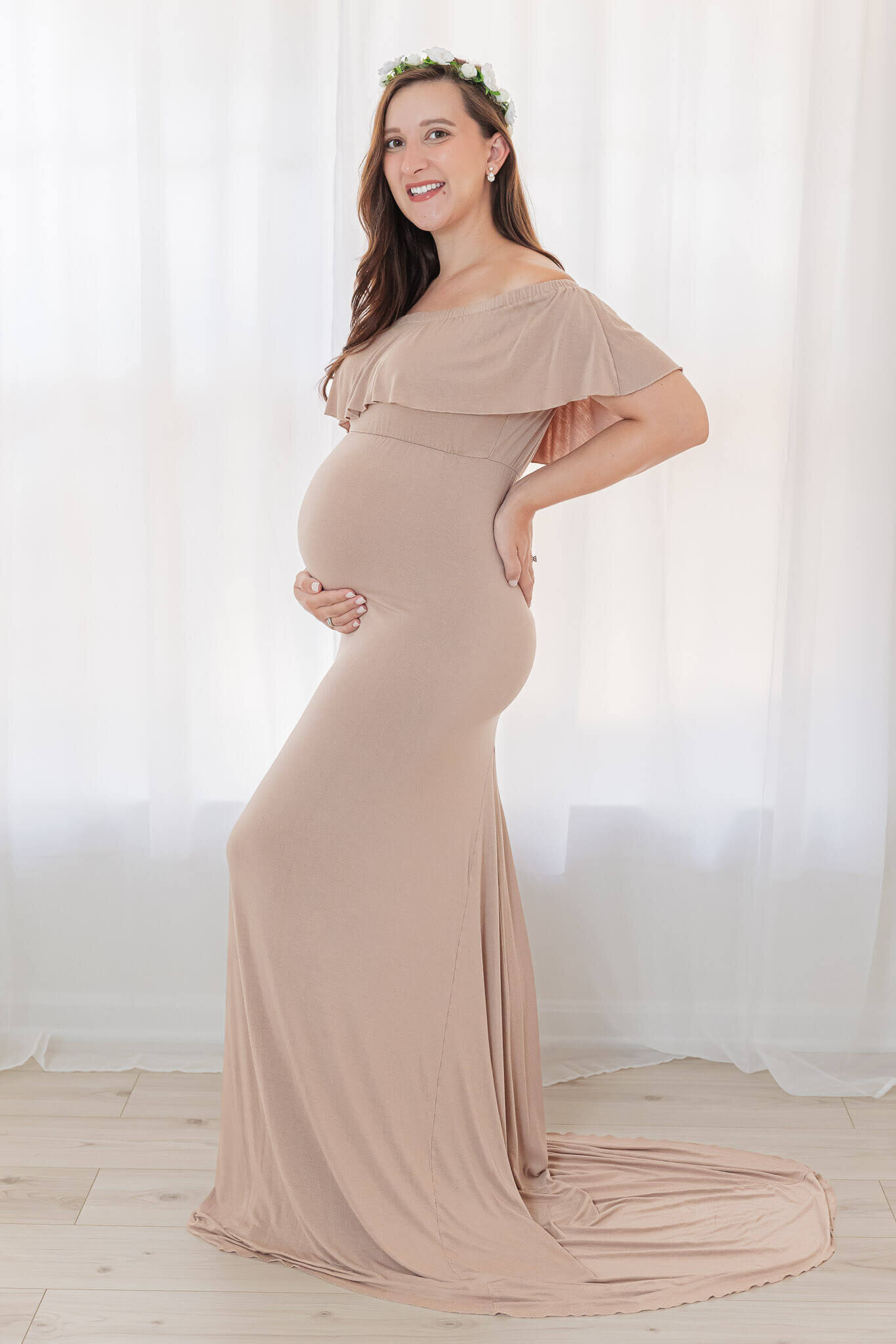 A mother-to-be in a tan dress posing in front of a window.