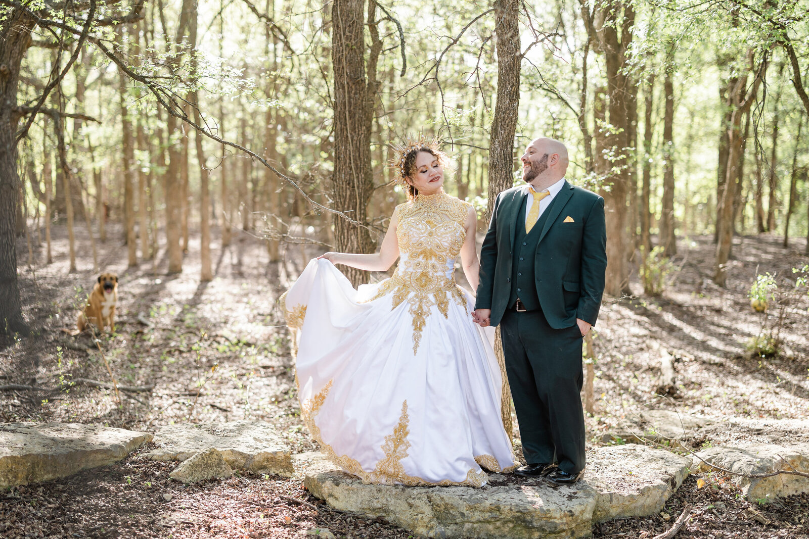 A bride in a gold crown and gold accented dress holds her groom's hand while fanning her skirt out. The groom is wearing a green suit. They are in a forest setting and standing on top of a rock. Their dog is in the background and out of focus. They are both smiling at each other.