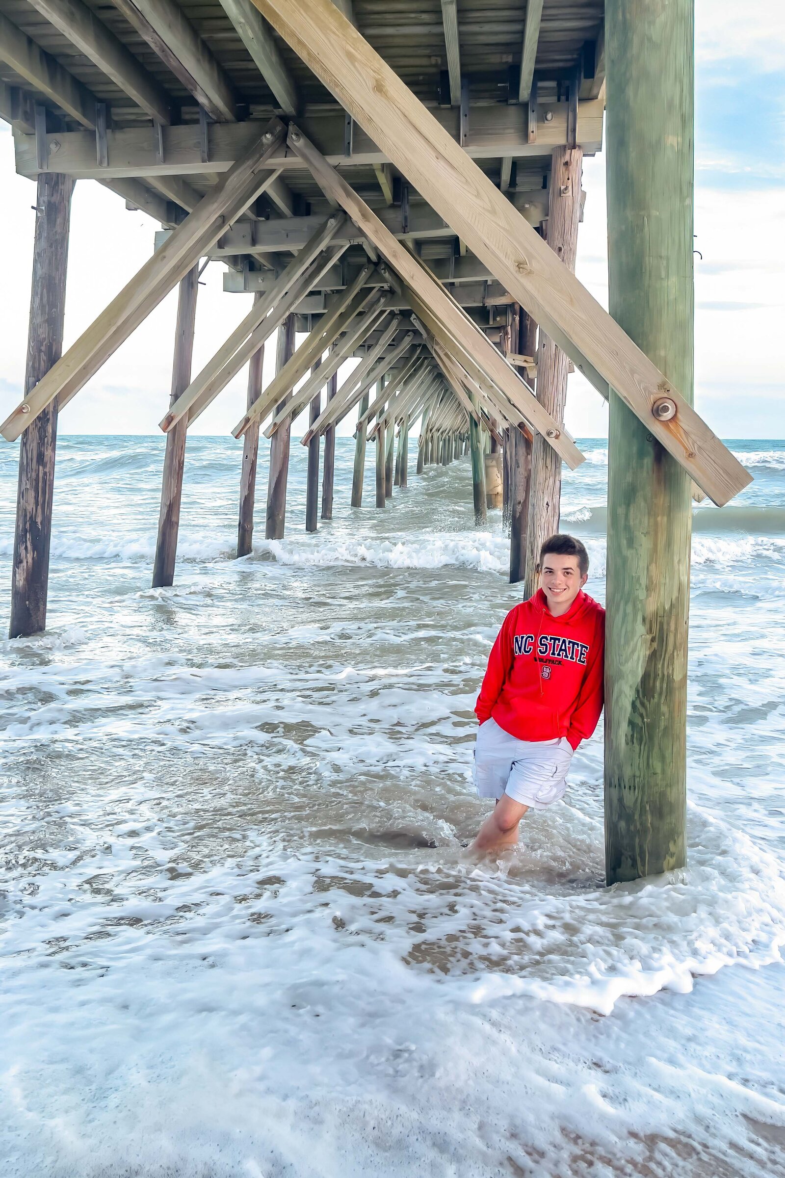 A high school senior wearing a red hoodie standing under a pier with waves crashing around in North Topsail Beach, North Carolina. This dynamic photo highlights the adventurous spirit of senior photography, ideal for capturing unique beach moments.