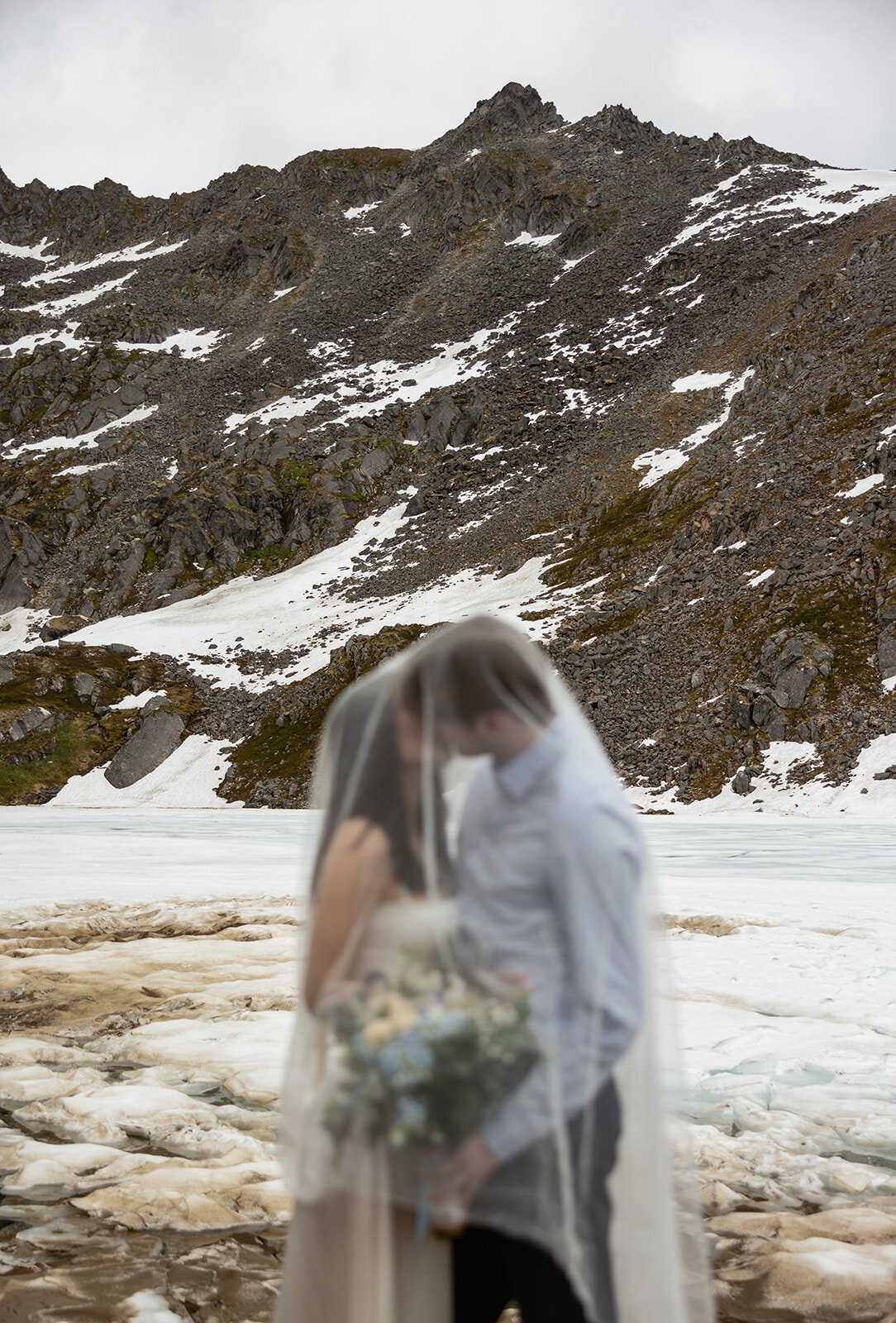 A couple shares an intimate moment beneath a bridal veil surrounded by snow patches and rugged mountain slopes during their Alaska elopement, captured by Sydney Breann Photography.