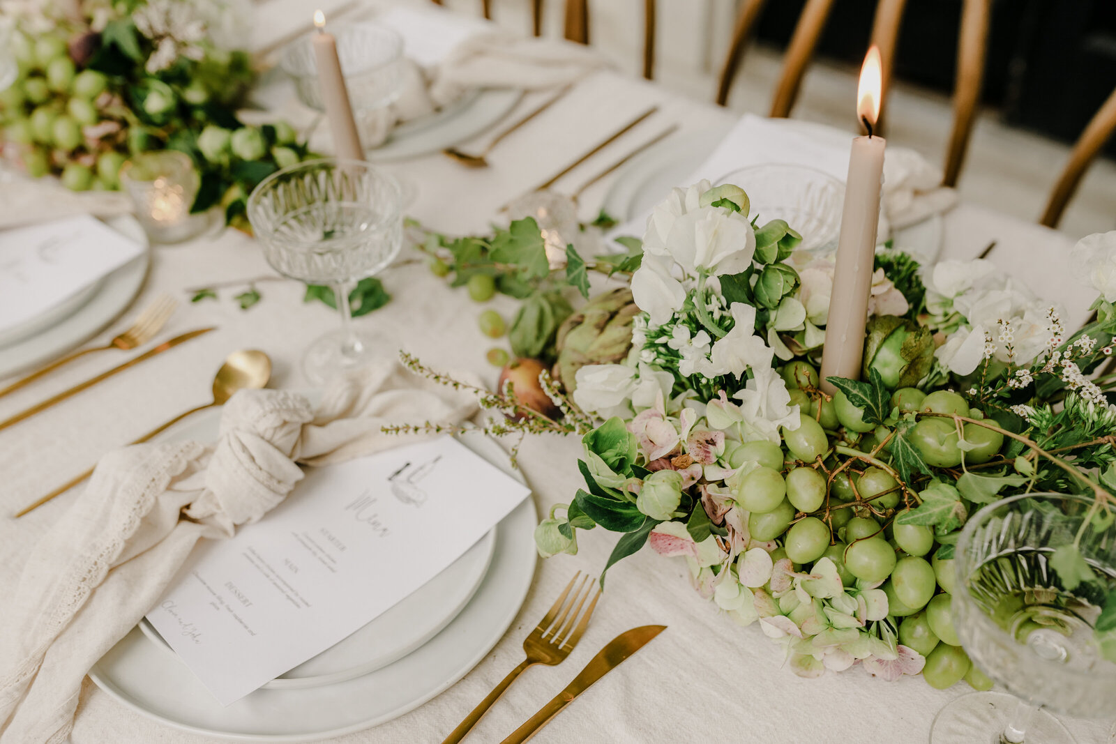 Editorial detail of a luxury wedding tablescape with soft florals, green grapes, taper candles, and linen textures styled with gold flatware and custom menus.