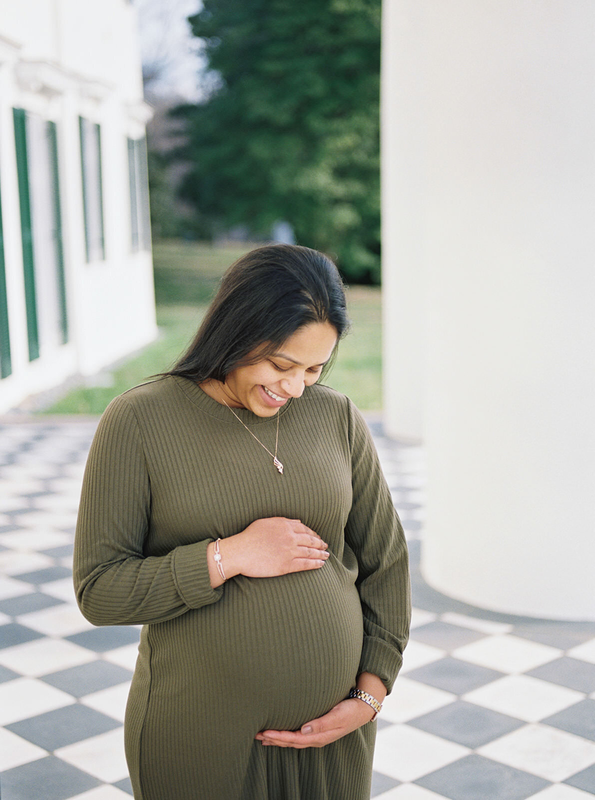Expectant mother in a green dress smiling warmly against a classic architectural backdrop.