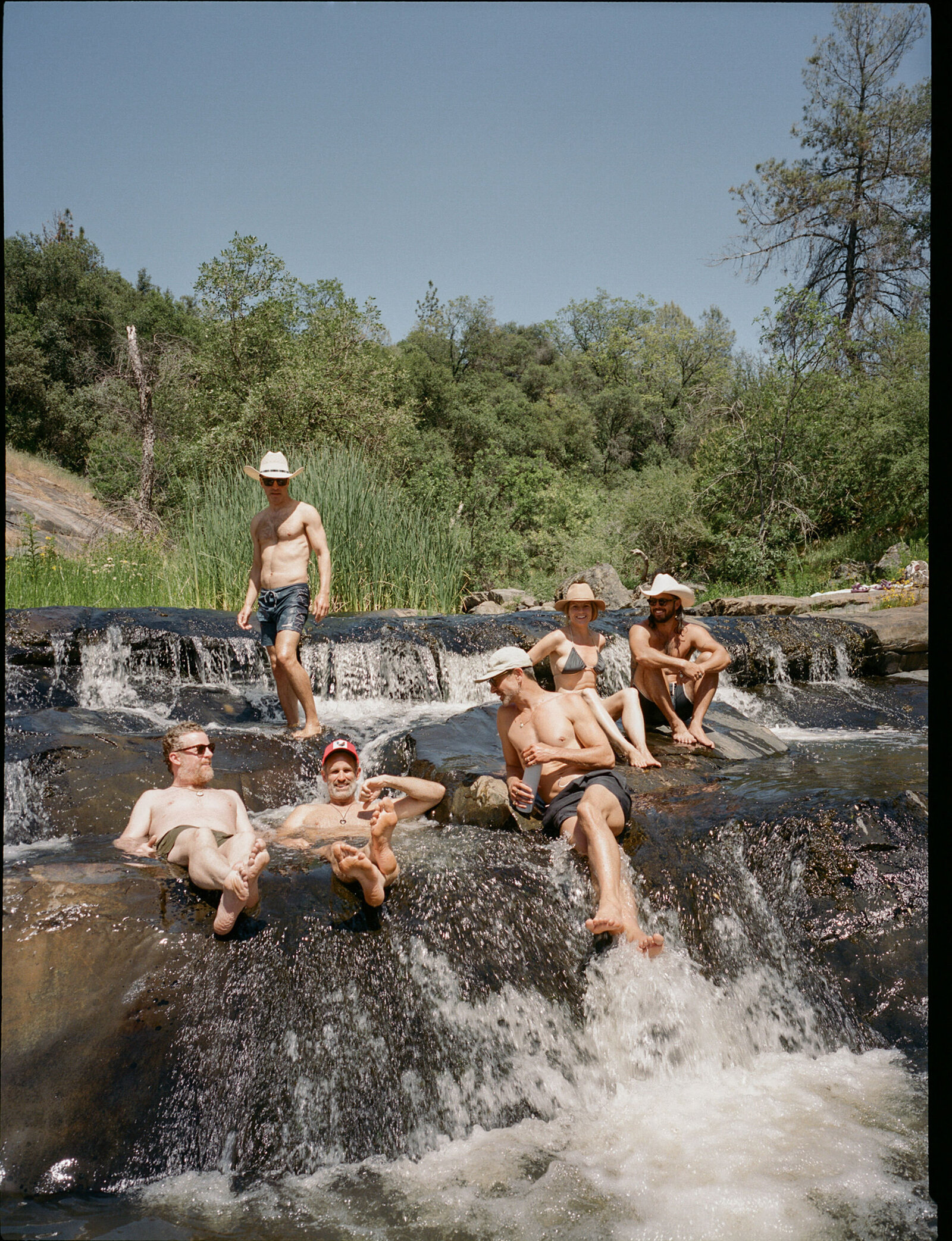 Group of friends in cowboy hats lounging on waterfall rocks in a river in the Sierra Nevada mountains during summertime