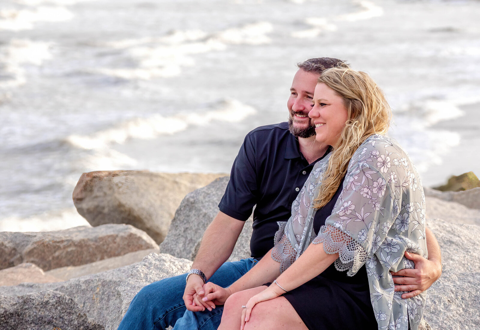 An engaged couple laughing and sitting together on large rocks by the beach in Wilmington, NC. This candid wedding photography showcases their genuine connection, ideal for military and healthcare professionals looking for unique wedding photography packages.