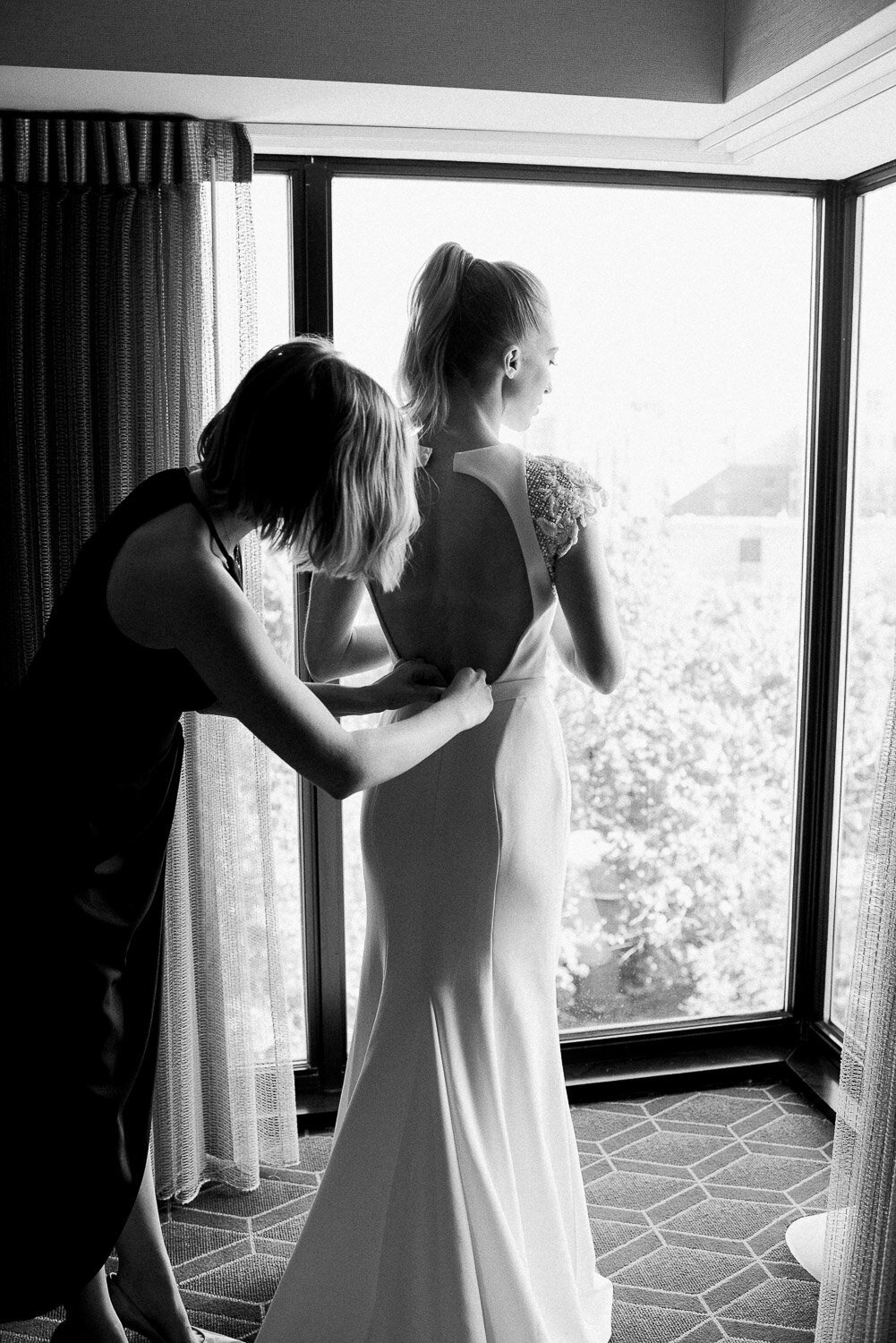 bride and bridesmaid button dress from behind in window