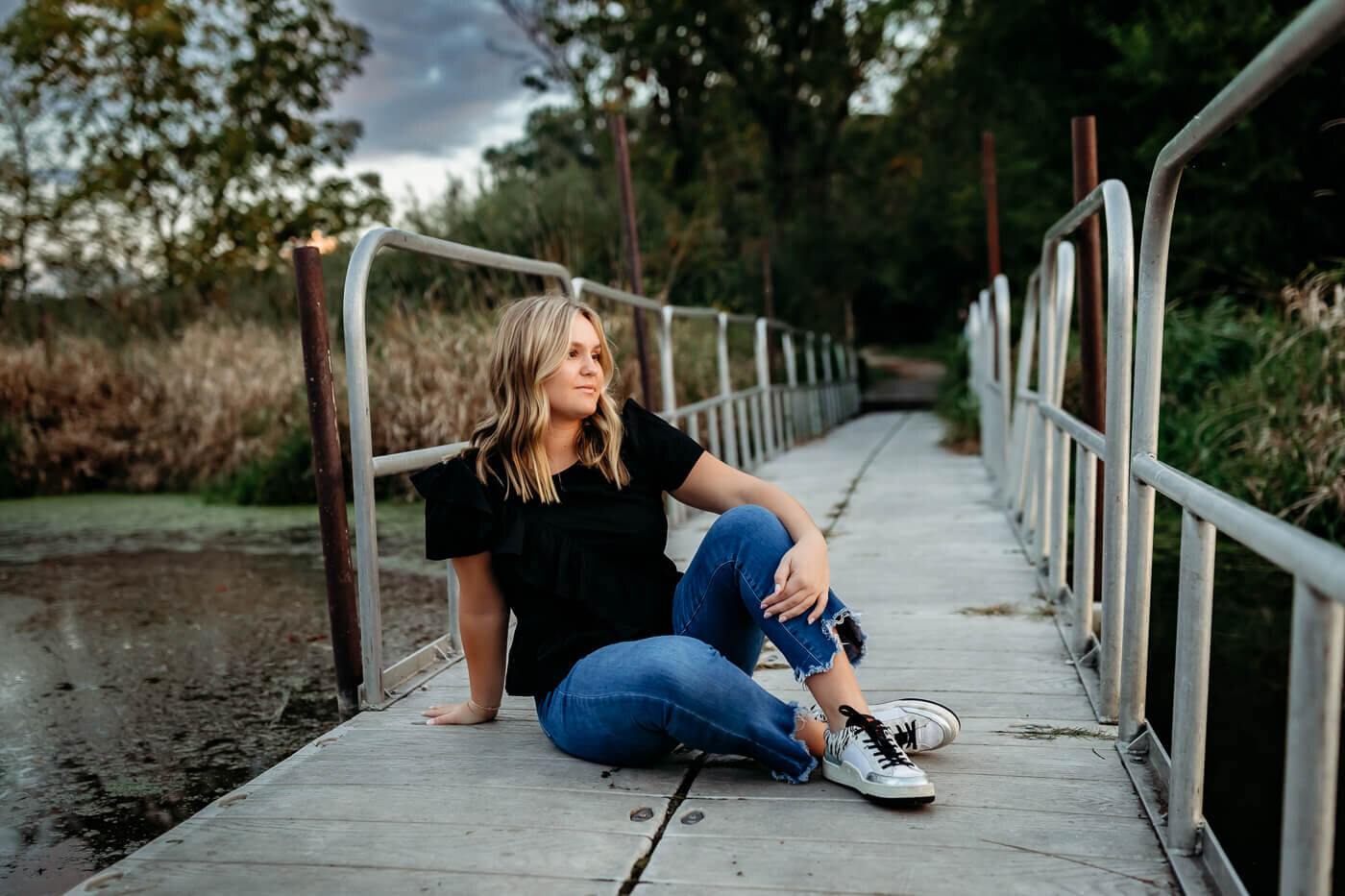 A woman with blonde hair sits casually on a wooden bridge. She is wearing a black top, ripped jeans, and black Converse sneakers. The bridge is surrounded by greenery, and the sky is cloudy.