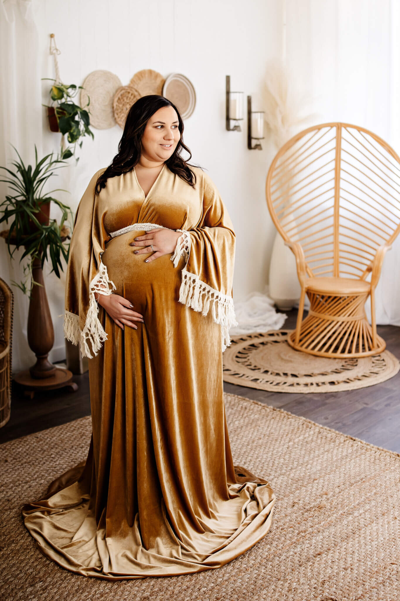A pregnant woman in a gold velvet dress stands on a woven rug, cradling her belly and smiling. The room features boho decor, plants, and a peacock chair—perfect for elegant Bloomington maternity photographer sessions or maternity portraits.