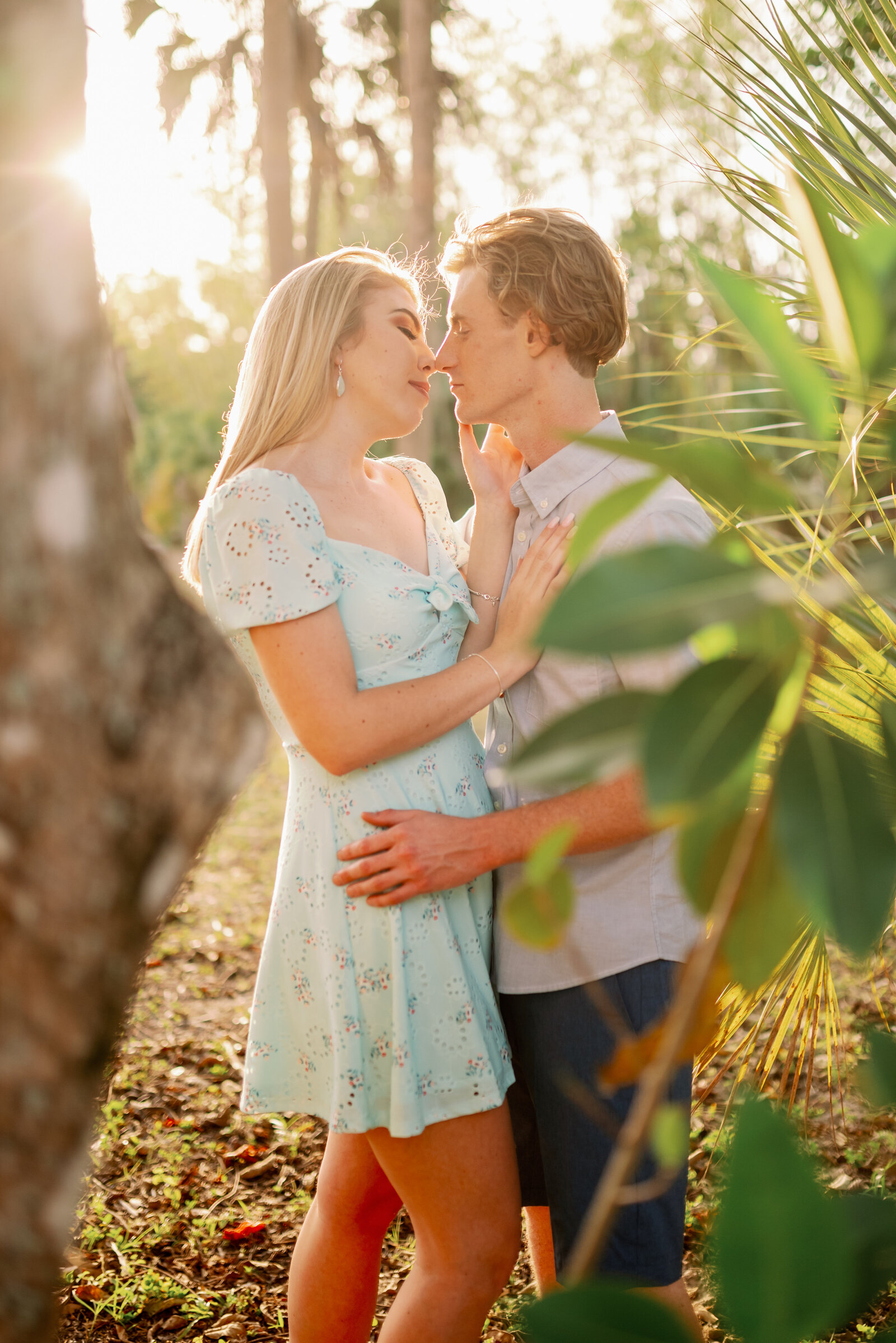 engaged couple about to share a passionate kiss with sunlight peeking through the trees behind them and leaves in the foreground
