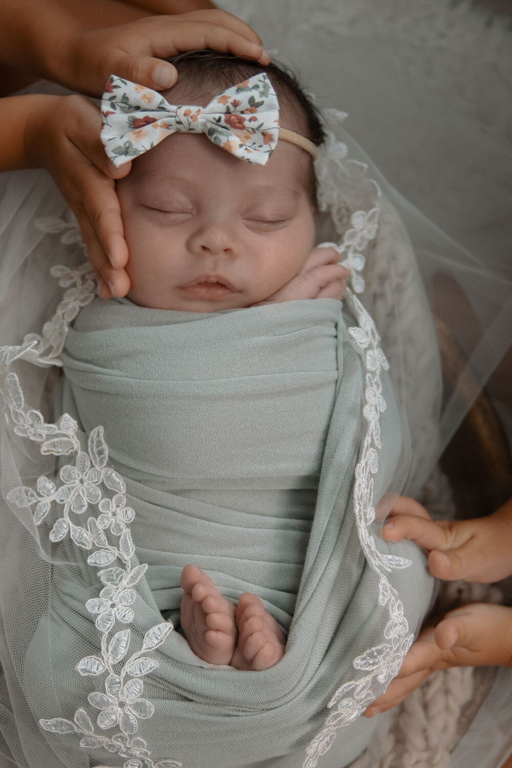 Newborn baby wrapped in green with floral lace and a bow, lovingly held by siblings