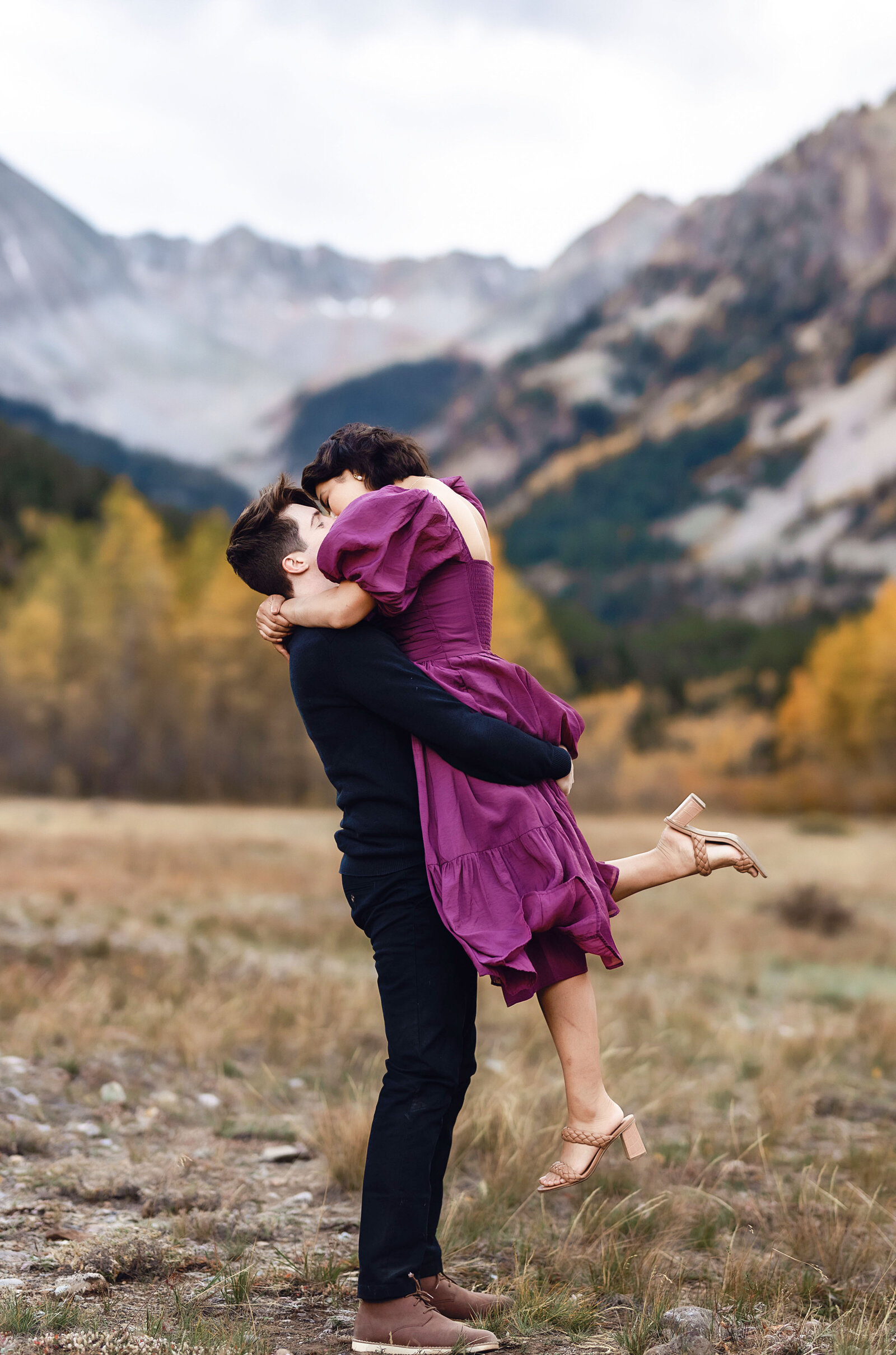 A man lifts up a. woman in a beautiful magenta dress and kisses her with a gorgeous Aspen mountain backdrop,