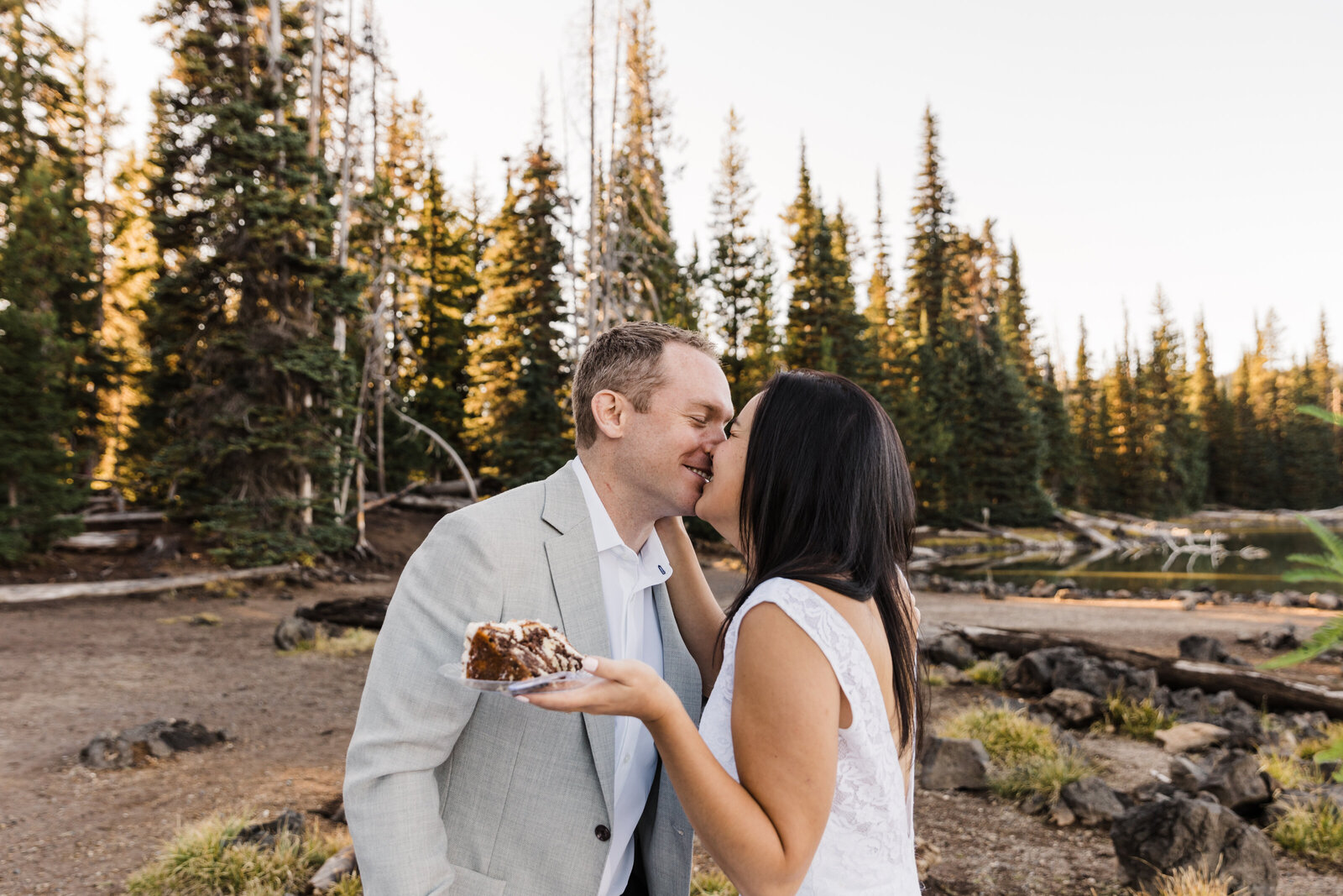 A bride, with long straight brown hair, holds a slice of cake on one hand while kissing her blond haired, grey jacket groom with the forest and Sparks Lake surrounding them at their Bend Oregon elopement. | Erica Swantek Photography