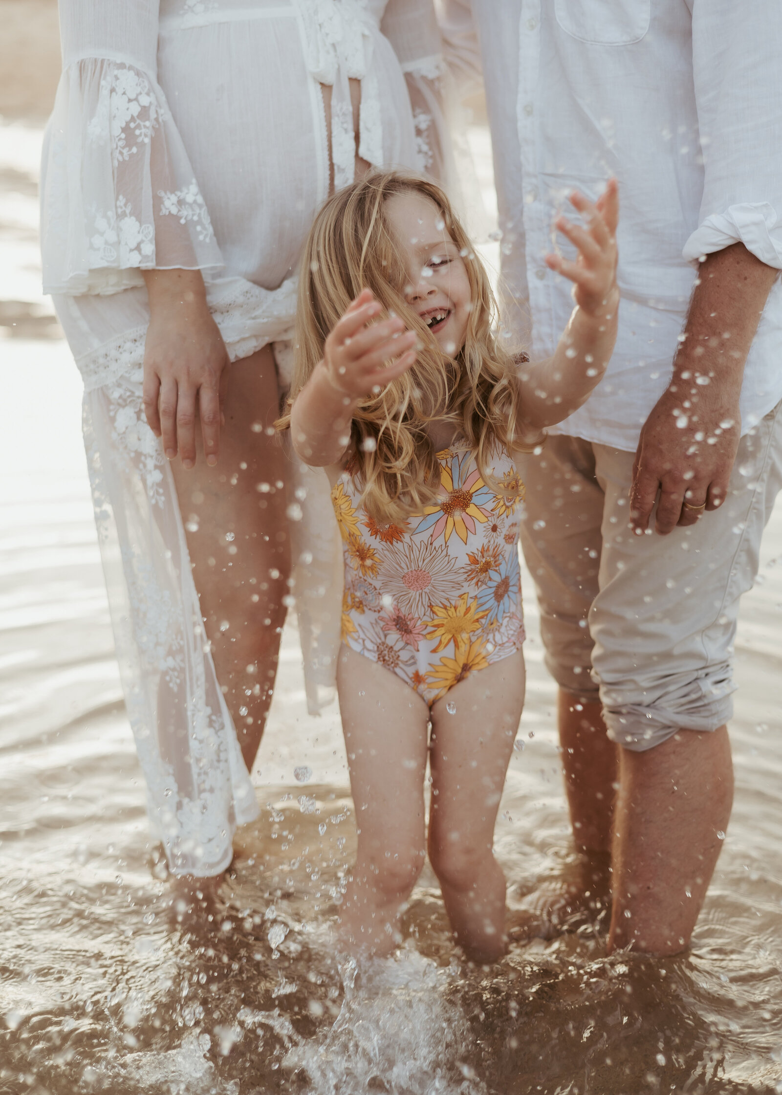 A close up photograph of a little girl splashing in the water with her parents behind her for their family photography experience at the beach.
