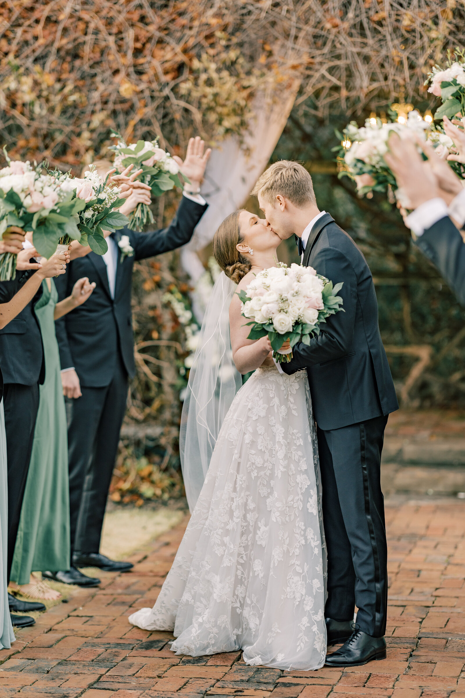 Bride and groom share a romantic kiss after walking through a tunnel of love created by their bridal party, celebrating their union with cheers and excitement.