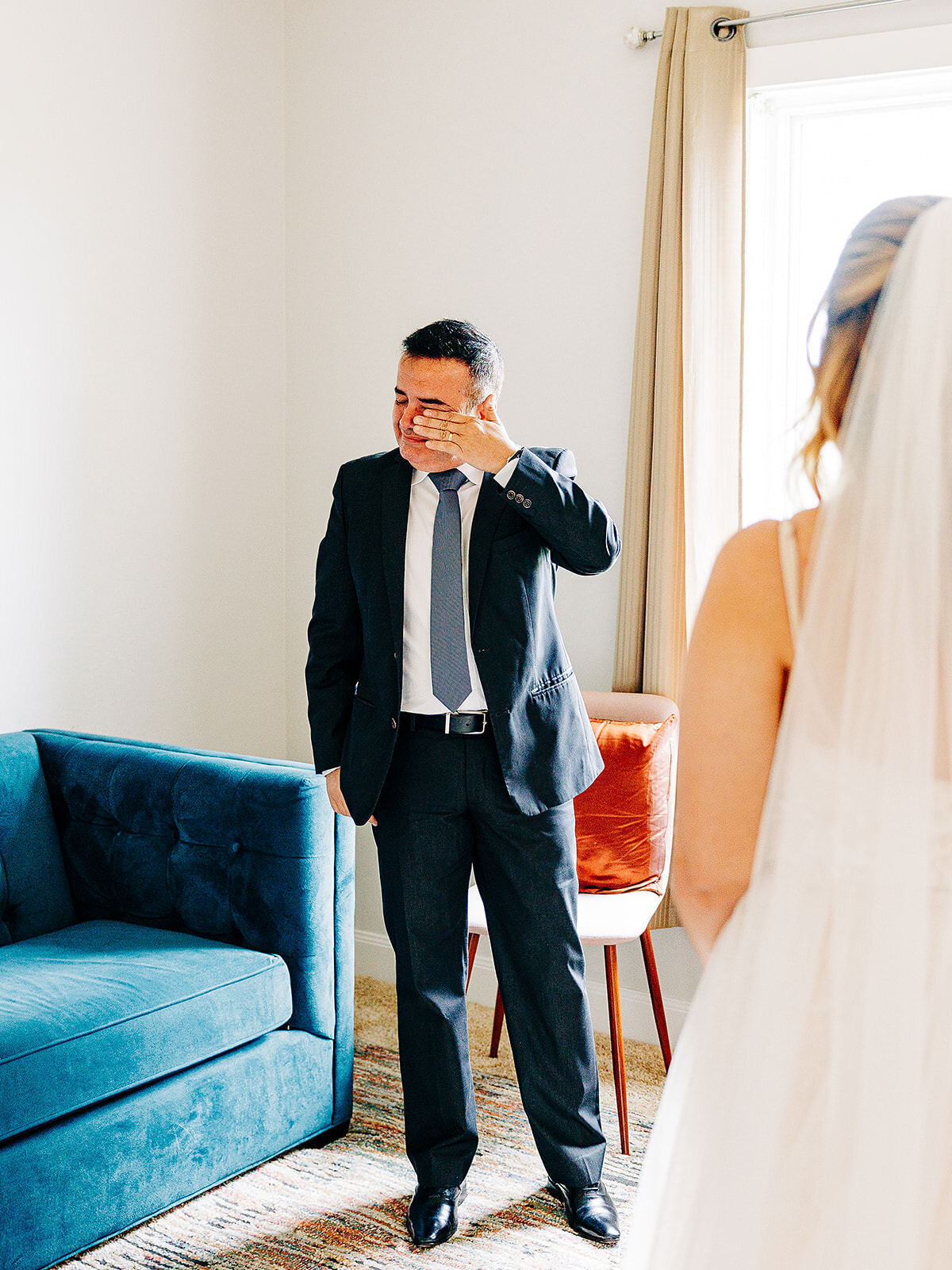 A man in a suit wipes tears from his eyes while standing in a bright room, looking toward a person in a wedding dress whose back is to the Austin wedding photographer.
