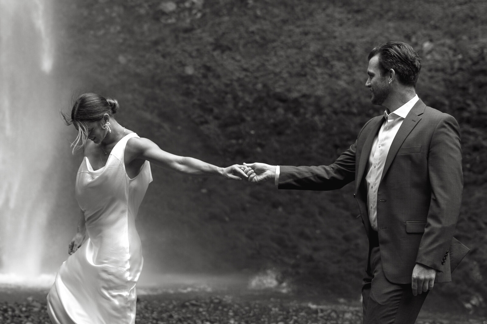 A bride and groom lead each other across the rocks in wedding attire at a waterfall in Nova Scotia. 