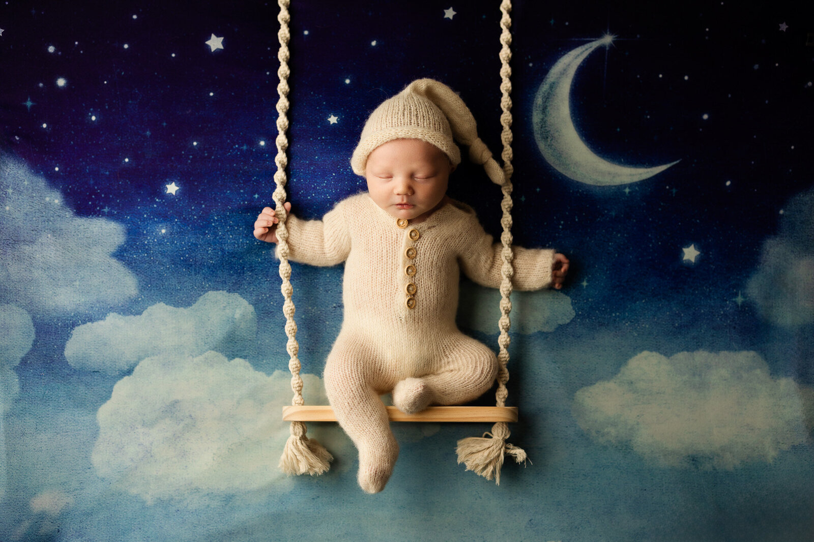 Baby boy in a cream knit sleeper and night cap, posed on a swing in front of a backdrop with the night sky.