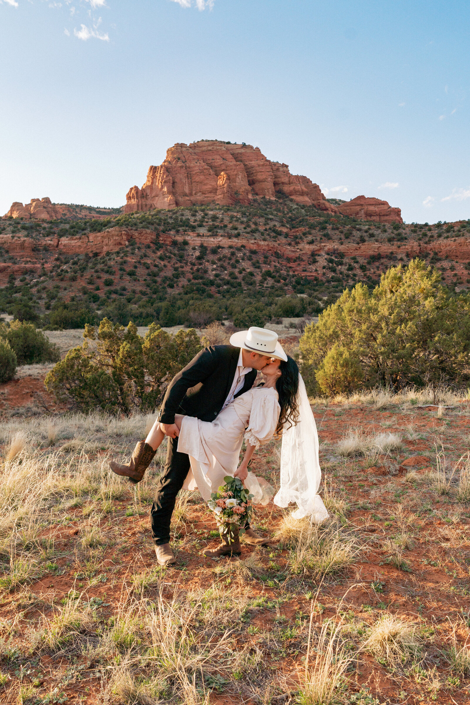 Western-inspired elopement at Bear Mountain in Sedona, Arizona featuring a groom in a cowboy hat dipping and kissing his bride as red rocks glow at golden hour.