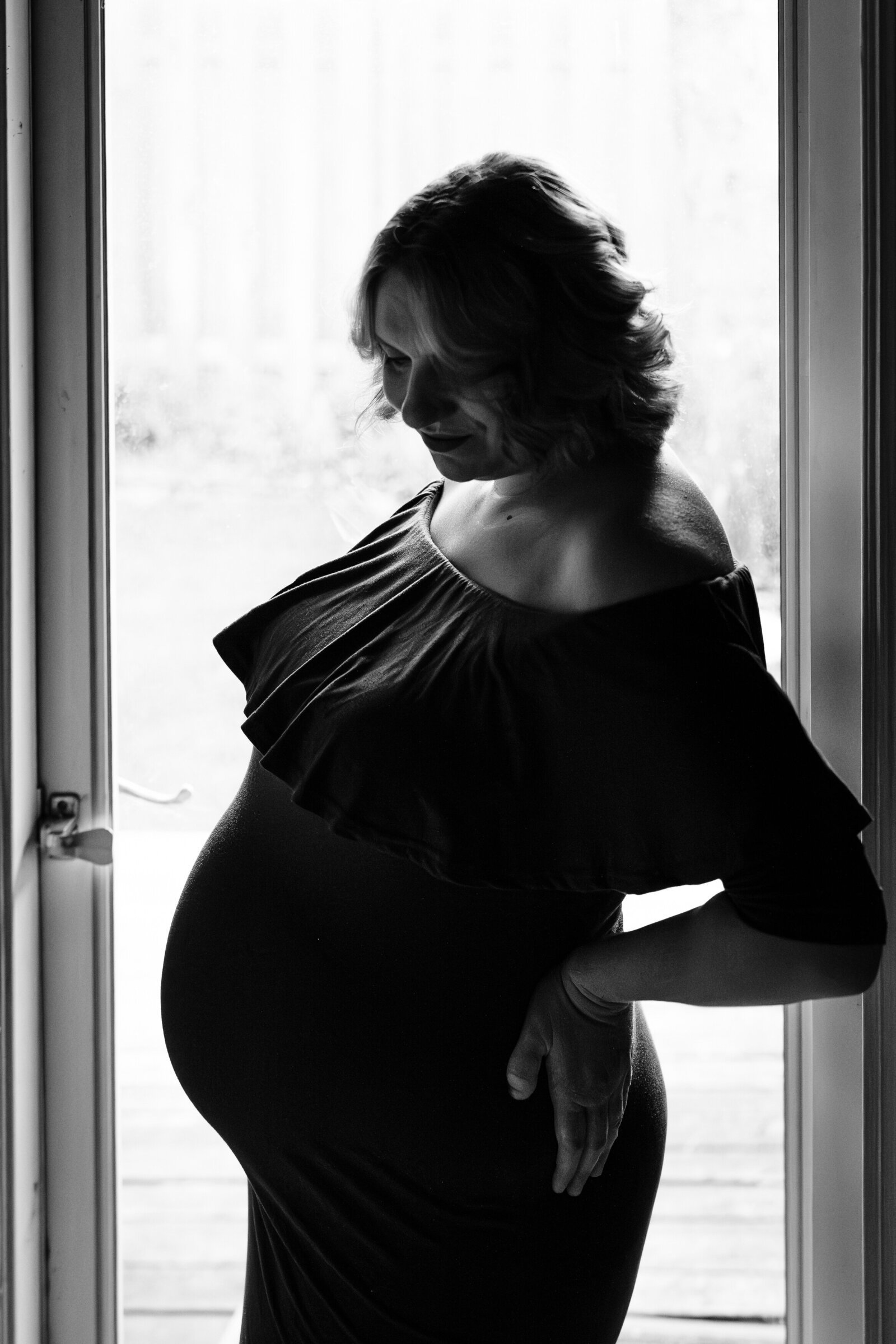 A black and white photo of a pregnant woman in a ruffled, off-the-shoulder dress, standing near a window with a soft smile. The light highlights her silhouette as she gently cradles her belly.