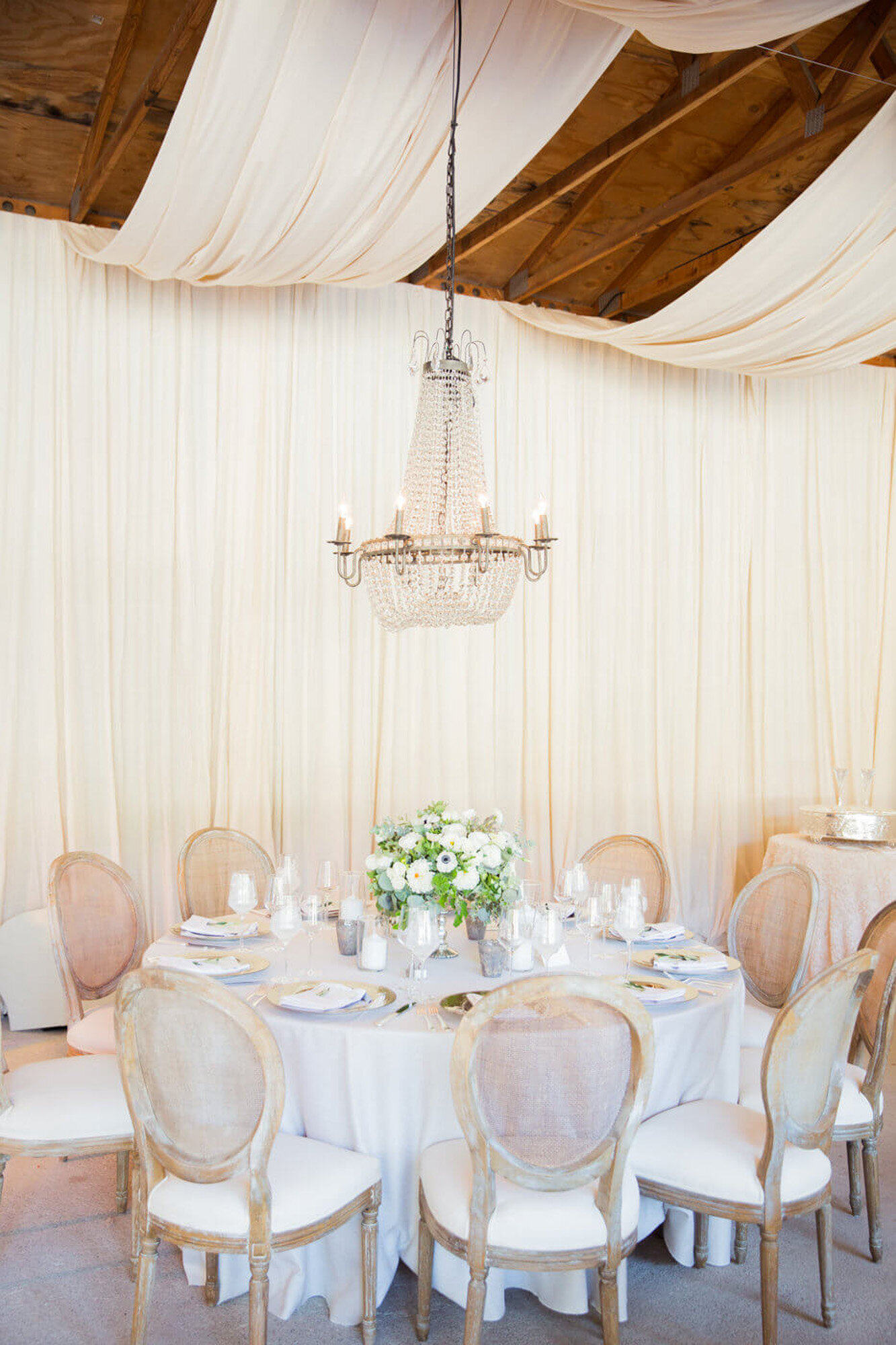 Luxurious round wedding table that has floral centerpiece, plates, utensils, glasses, candles. and a chandelier