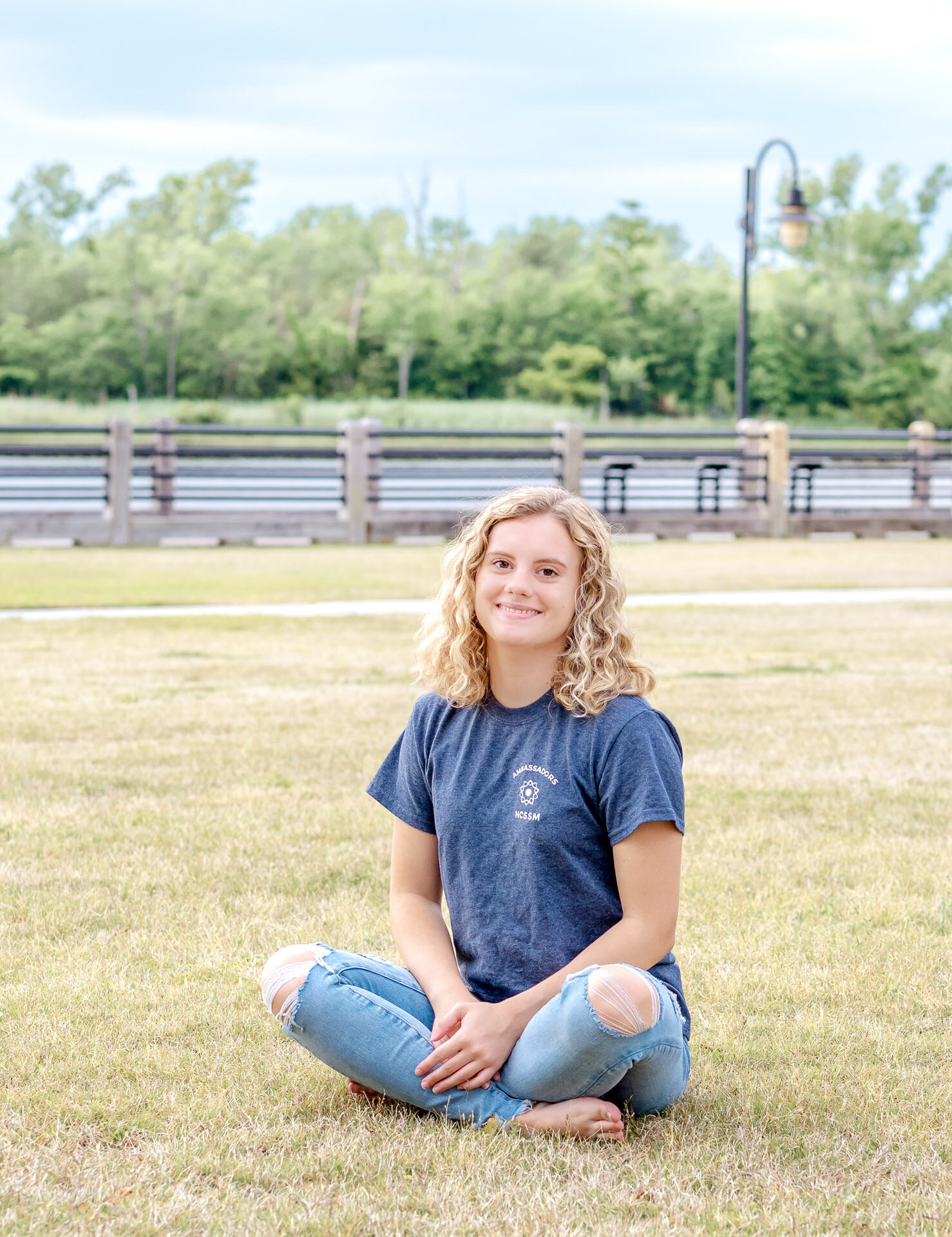 A high school senior sitting casually on the grass by the Cape Fear River in Wilmington, North Carolina, wearing a blue shirt and jeans. This relaxed image highlights the charm of natural senior photography, ideal for capturing youthful spirit.