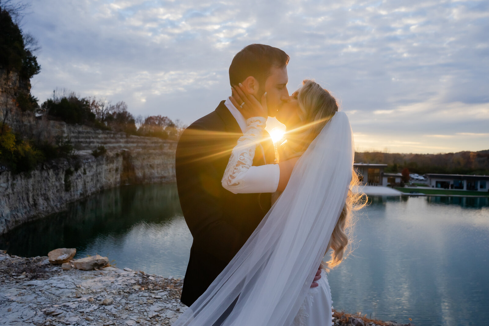 Bride and groom standing at sunset bluffs. They are kissing and her ring hand is holding his face. She has a long veil and gloves, he is in a black suit. The sunset is perfectly centered between them.