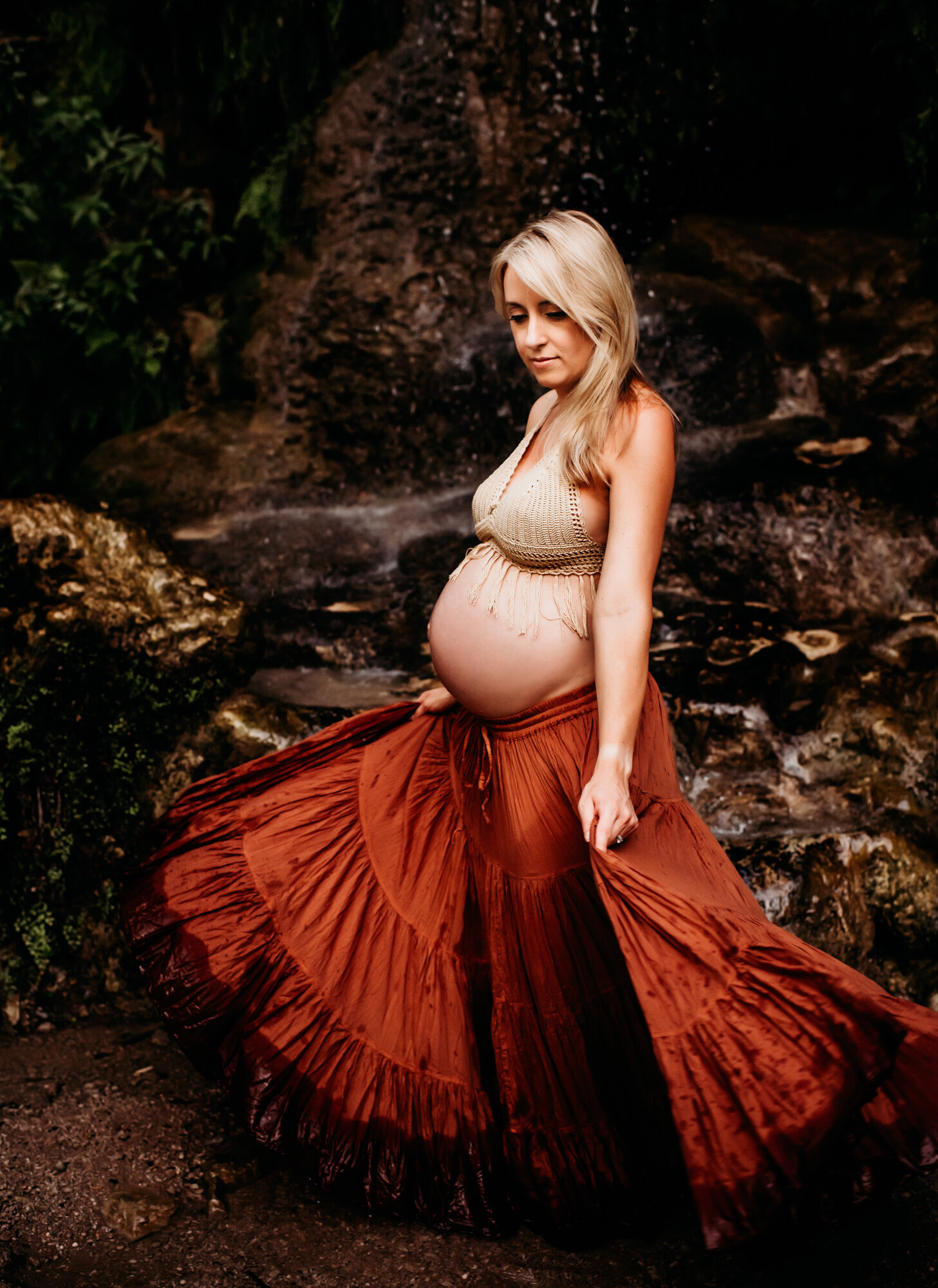 Maternity Photographer, A pregnant woman holds her long skirt as she stands near forest streams