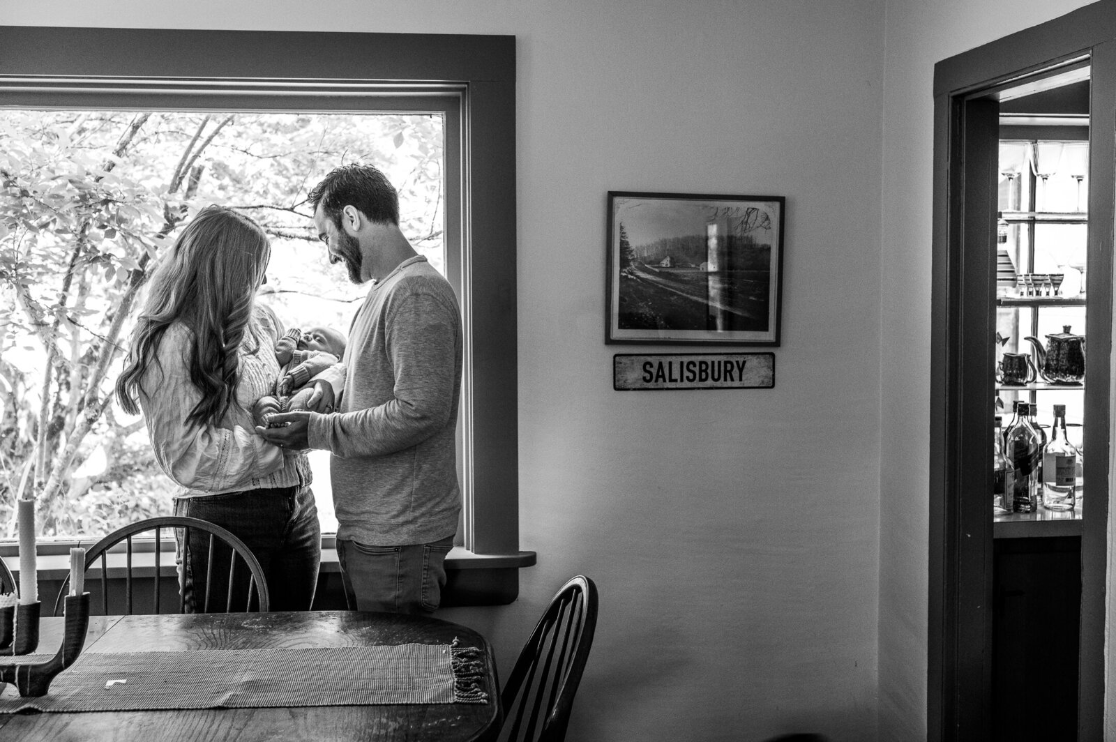 Mother engaging with her partner as he lifts their baby by a sunny window, in a candid black and white frame