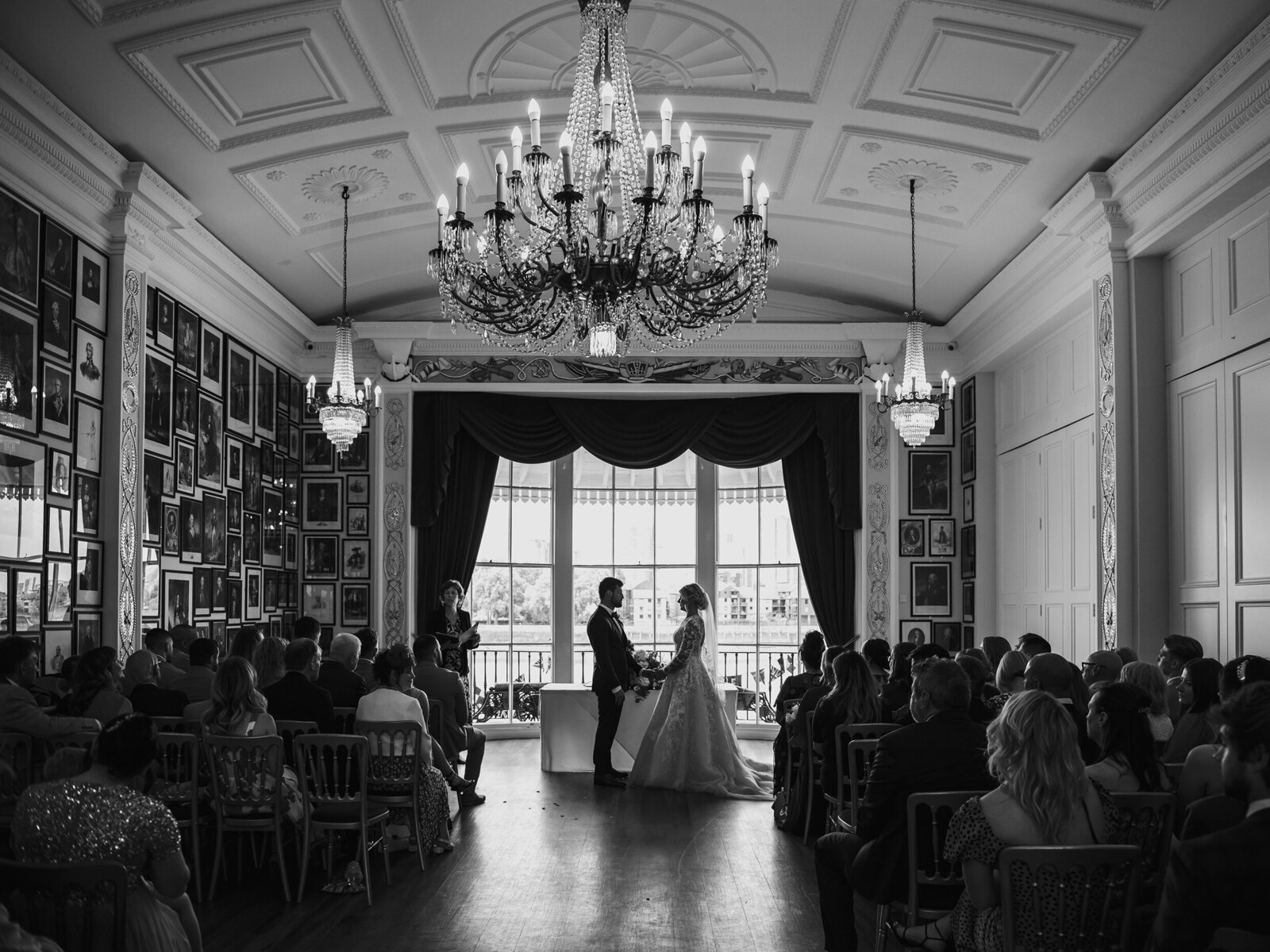 Black and white photo of a wedding ceremony in an ornate room with chandeliers as the couple stands at the altar