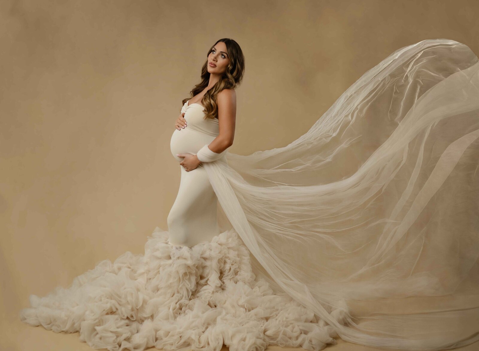 maternity photo of Mom in cream gown with tulle tossed in air behind her and her holing her baby bump. Photographed at Atlanta maternity photo studio.