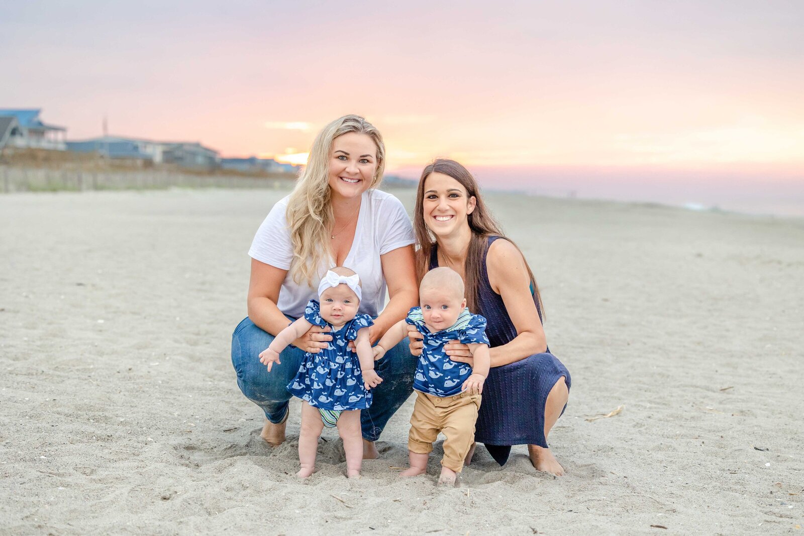 Two mothers hold their twin babies on the sandy beach at sunset, dressed in coordinating outfits. The colorful sky adds warmth to this tender family moment. Perfect for professional family photography in NC, capturing intimate seaside interactions.