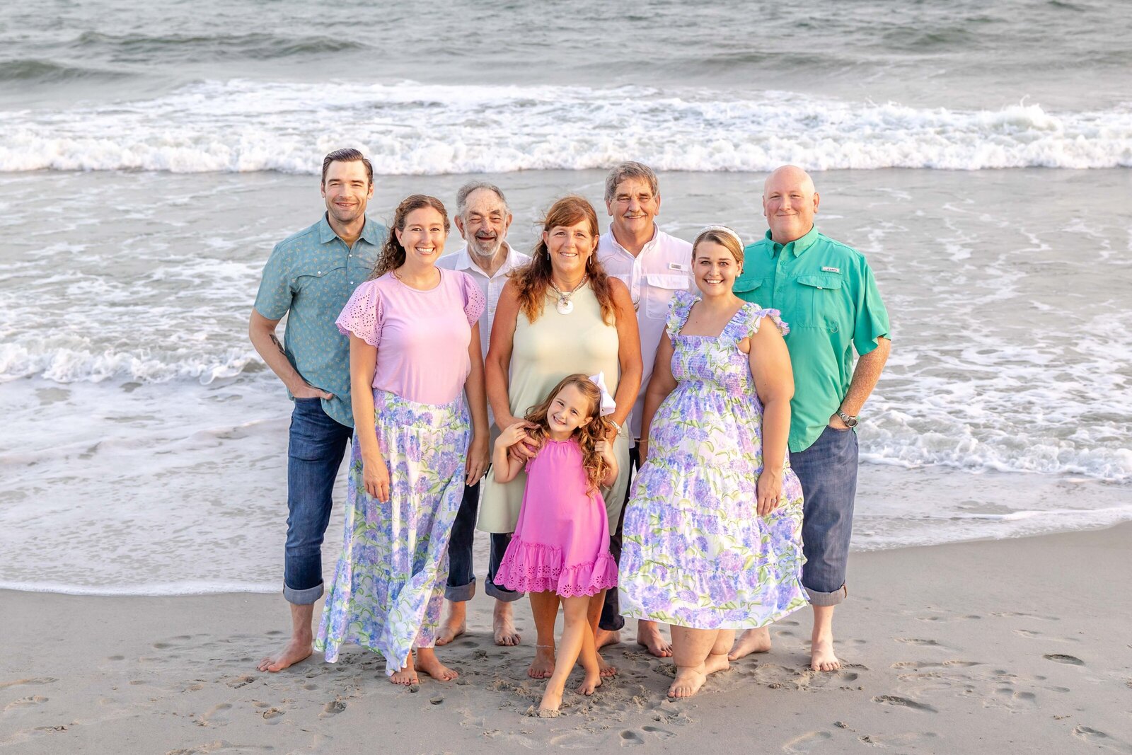 A family poses barefoot on the beach, dressed in vibrant pastel outfits with floral patterns. The ocean waves and setting sun create a warm and inviting atmosphere, ideal for professional family photography in NC, showcasing joyful seaside moments.