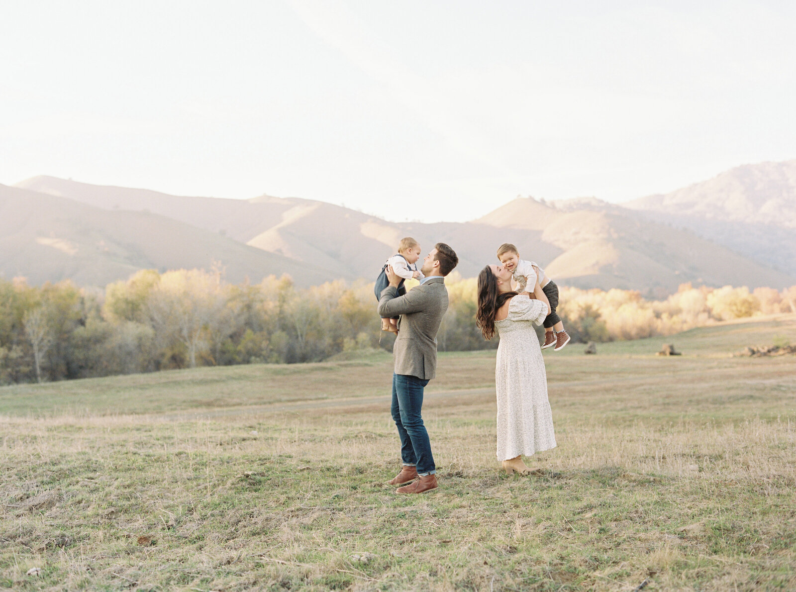 family outside in a field mom and dad picking up their sons and kissing their cheeks