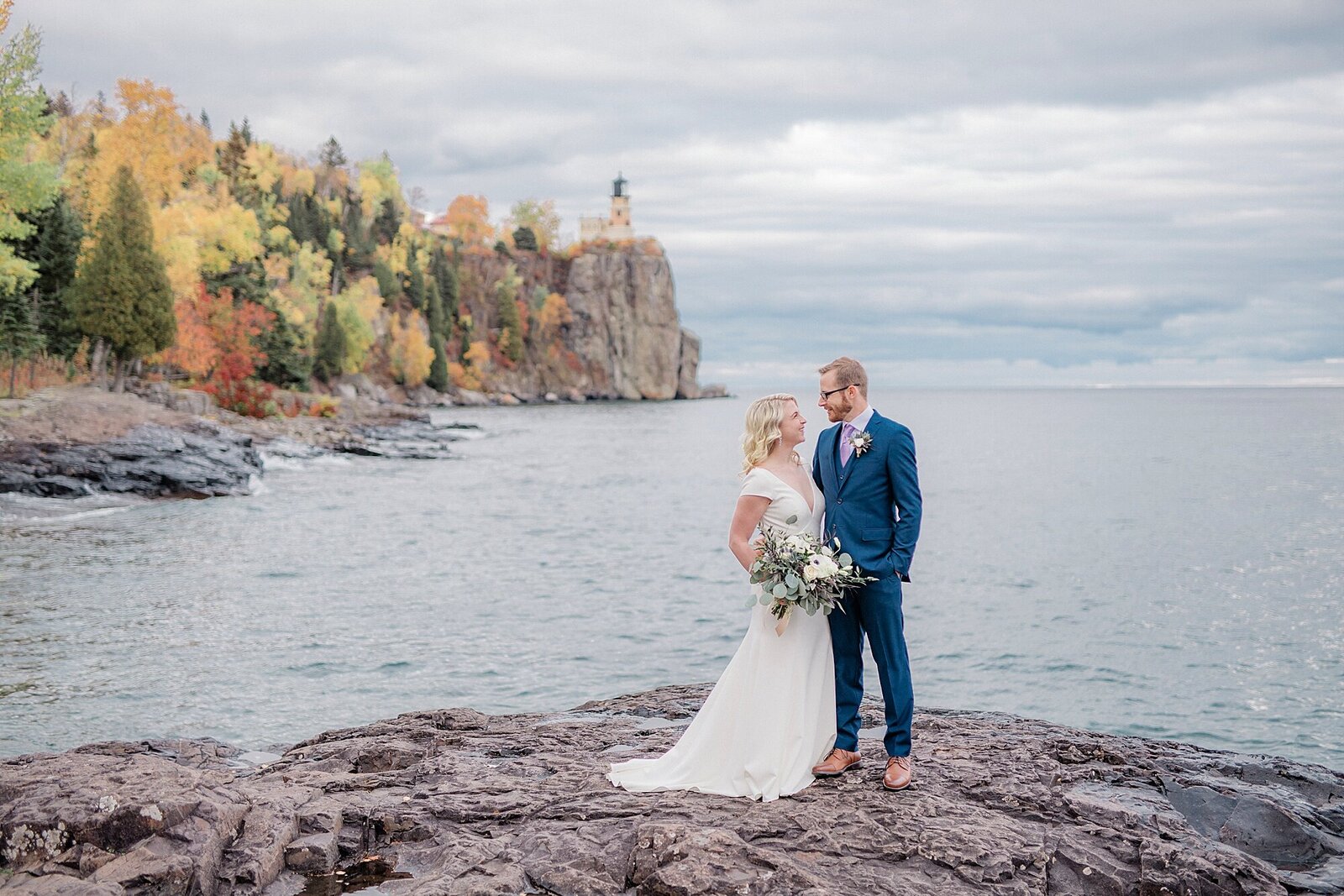 A couple smile at each other along the shores of Lake Superior with  Split Rock Light House in the background of their Minnesota wedding.