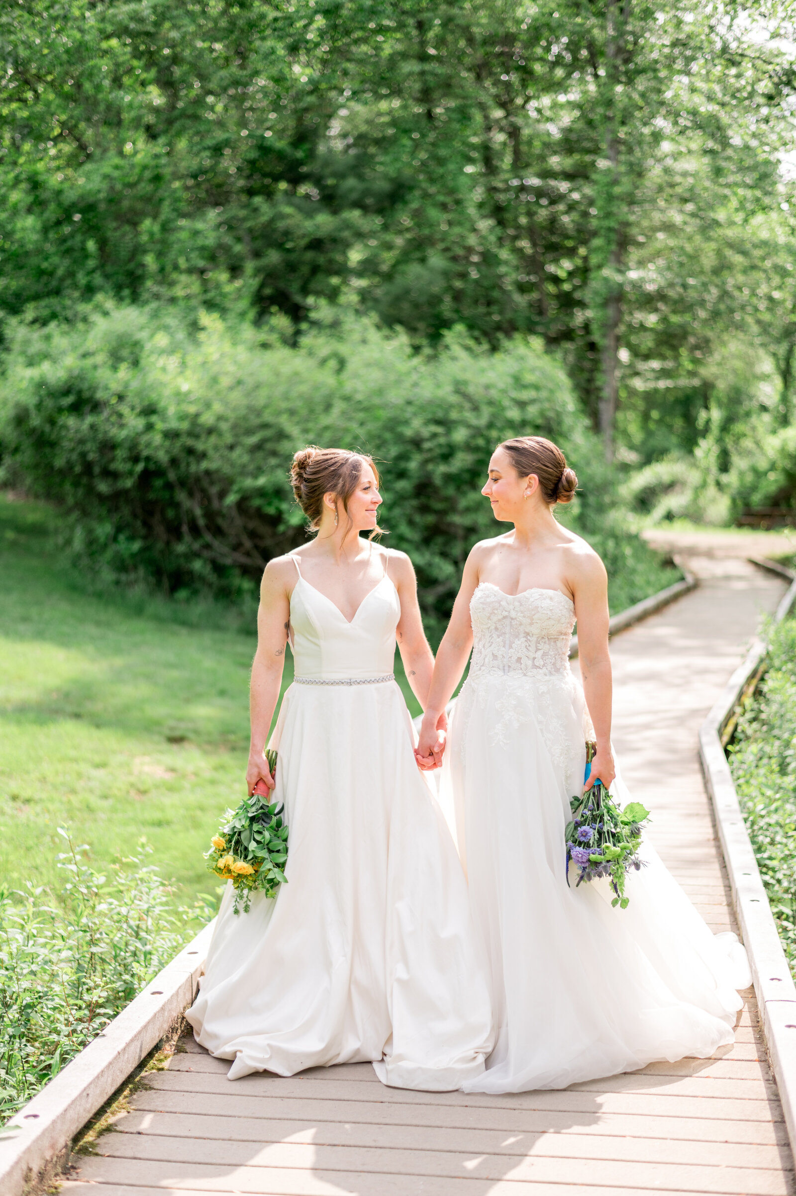 Two brides holding hands and smiling at each other on a sunlit path surrounded by lush greenery, both in flowing white gowns with vibrant spring bouquets — romantic, bright, and joy-filled.