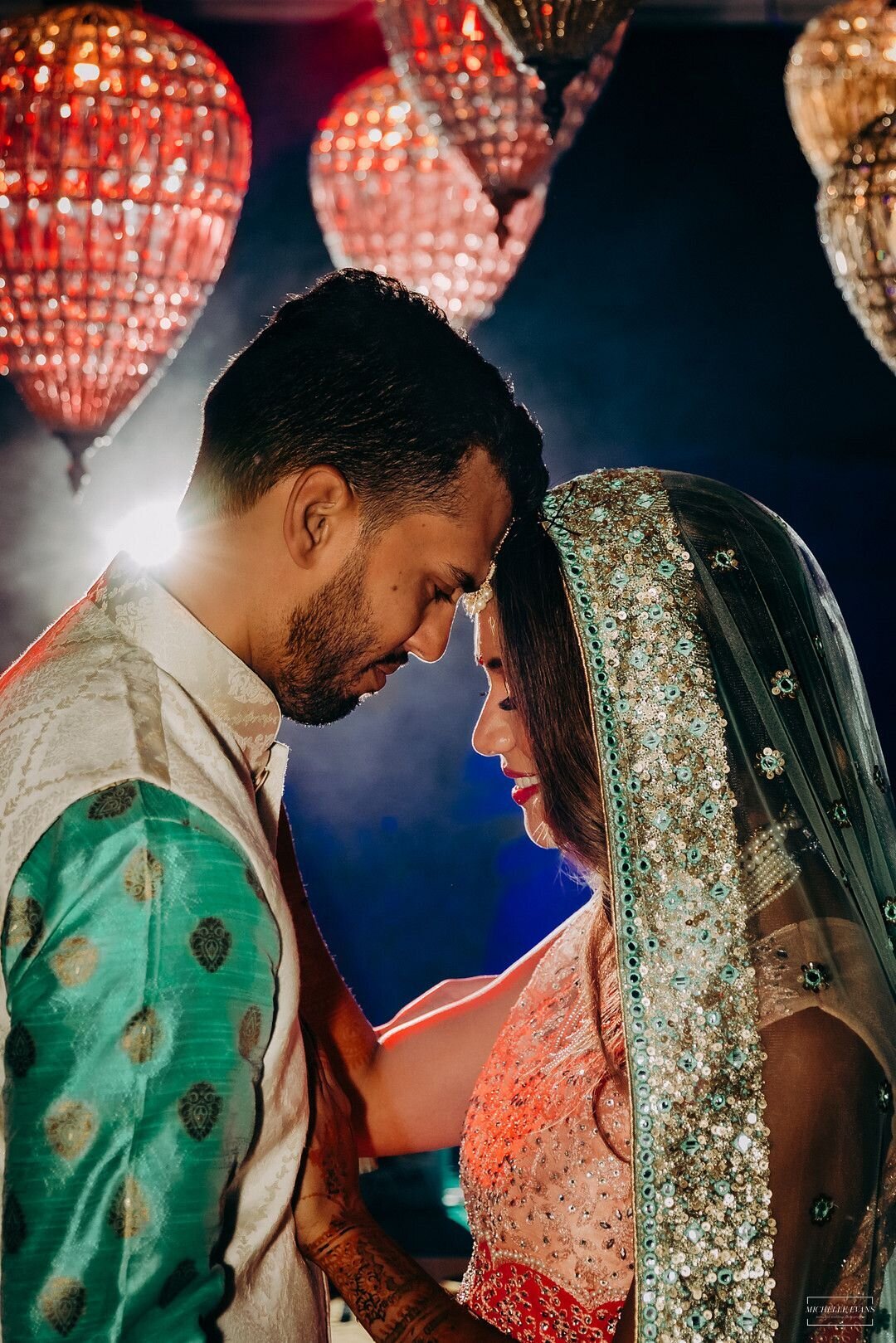 Romantic South Asian wedding portrait with couple in traditional attire under glowing chandeliers, featuring intricate embroidery, mehndi, and dramatic lighting—captured at a Memphis celebration styled by Wells Design Co.