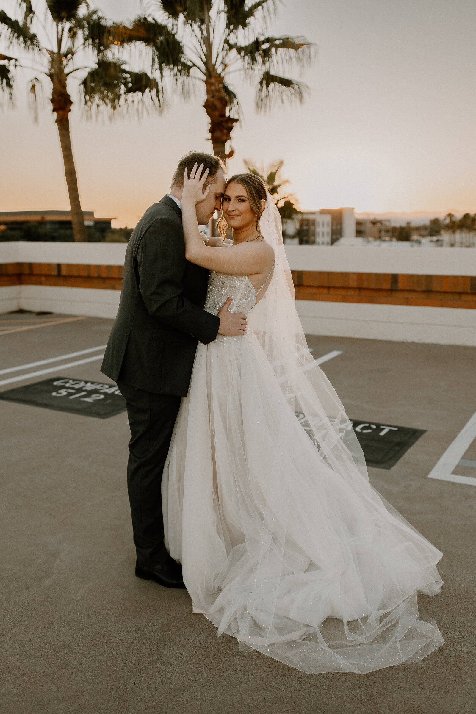 A bride and groom with their arms around each other standing in a parking lot.