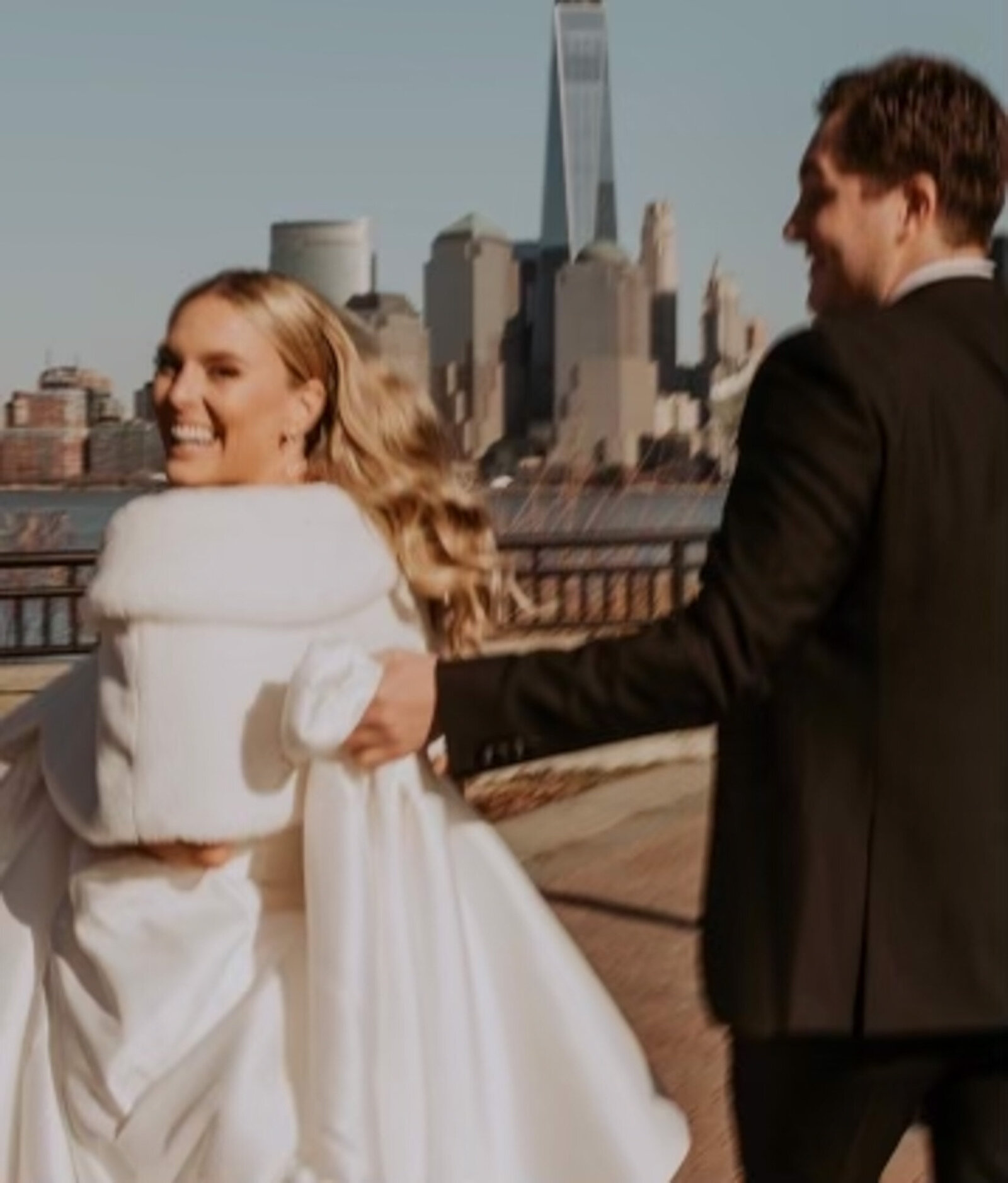 bride smiling and wearing a white dress and a groom in a black suit walk hand in hand along a waterfront promenade